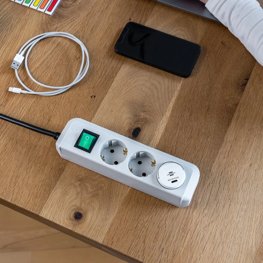 Weiße USB-C Steckdosenadapter-Mehrfachsteckdose mit zwei Schuko-Dosen und grünem Ein/Aus-Schalter auf Holztisch, Smartphone liegt daneben