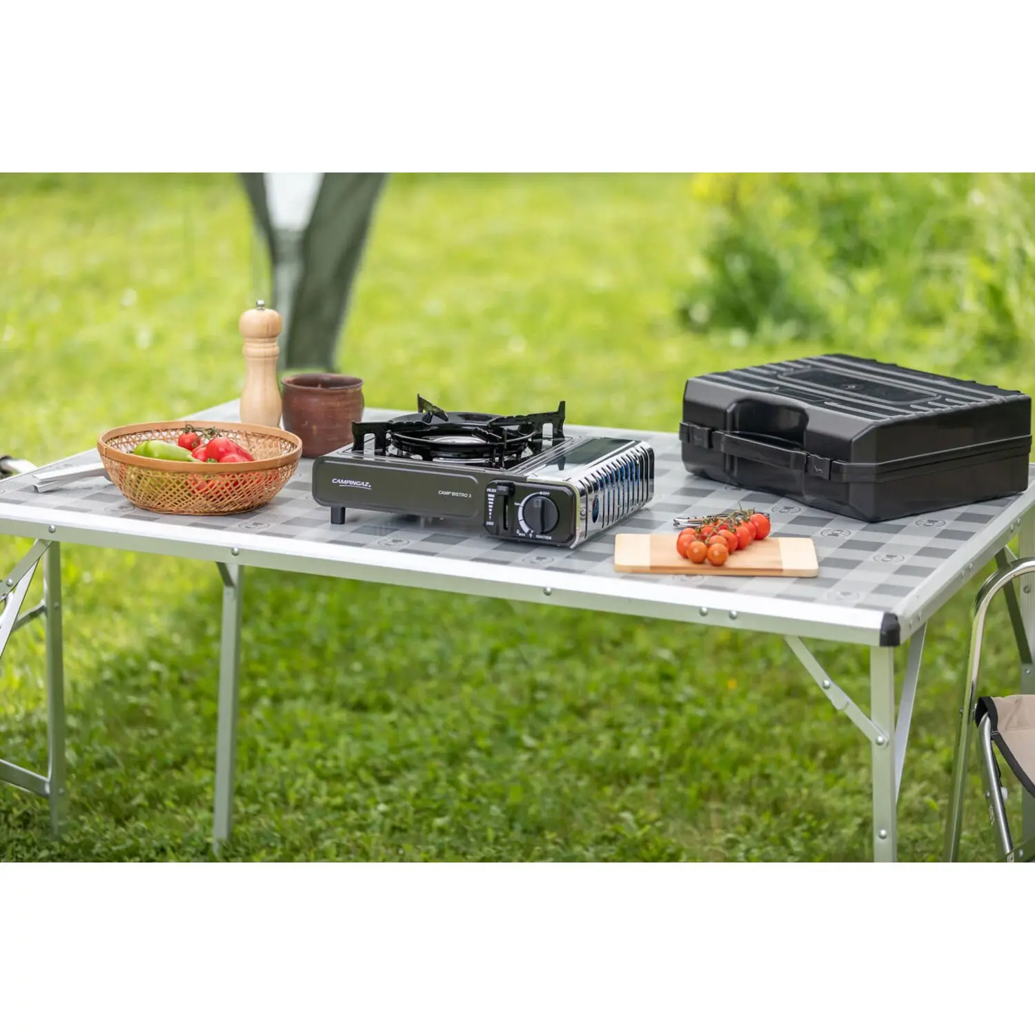 Auf einem Camping-Tisch steht der Camp’Bistro 3 Gaskocher mit zwei Brennern, daneben eine schwarze Transportbox, Schneidebrett mit Tomaten