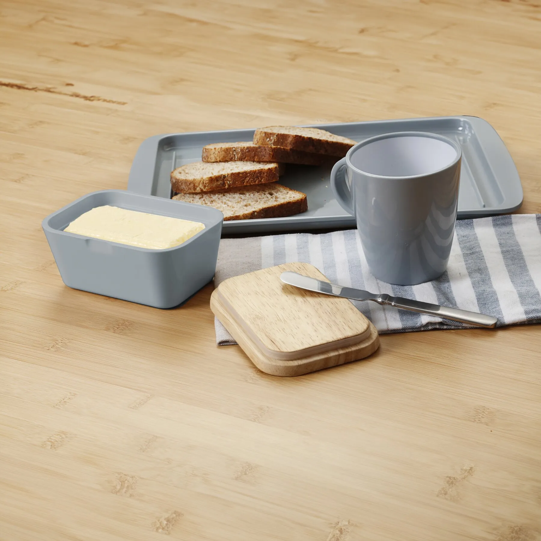 Graue Butterdose mit geöffnetem Deckel und Butterblock, neben Brotscheiben, Tasse und Buttermesser auf einem Frühstückstablettાન્ય
