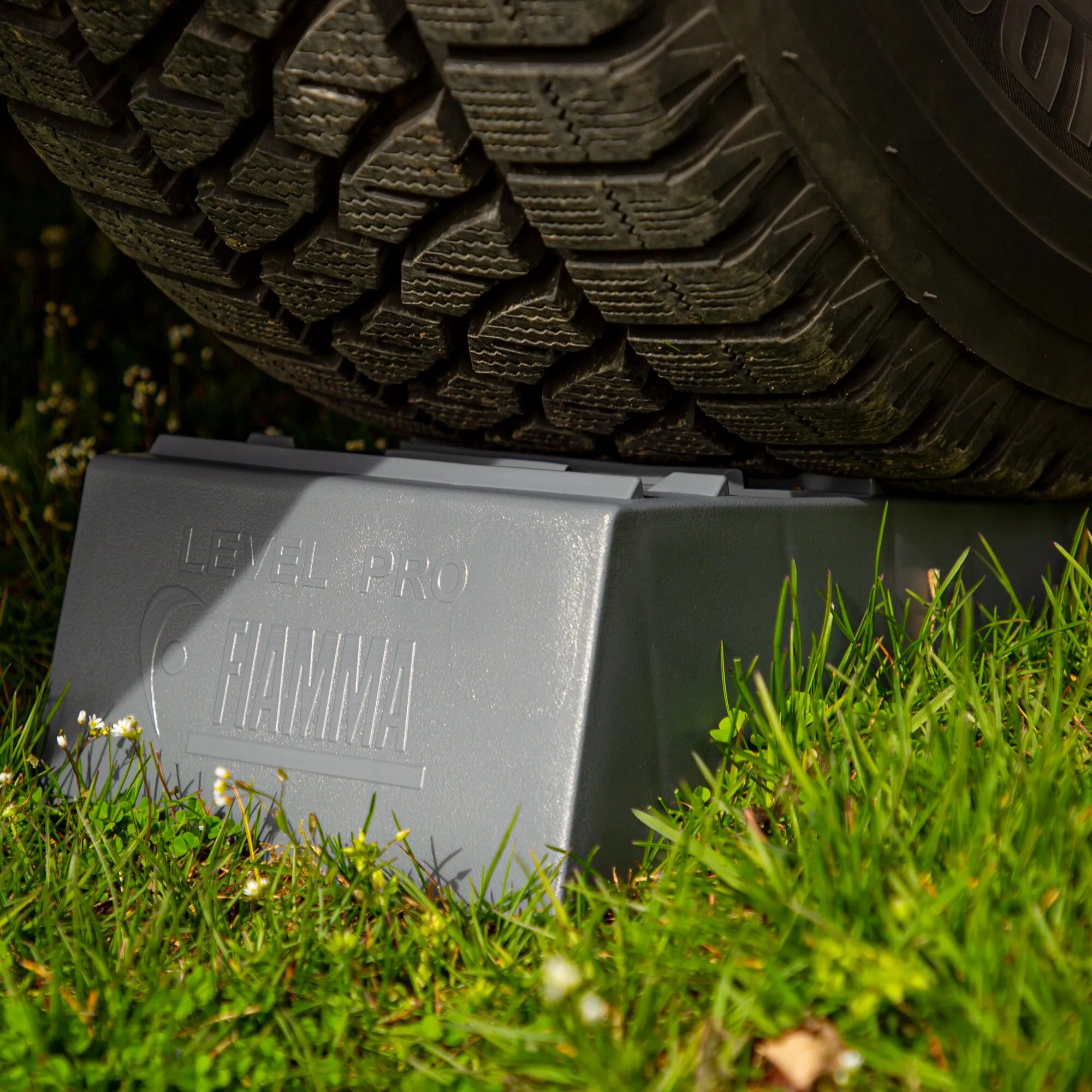 Grauer Auffahrkeil Level Pro im Gras neben dem grobstolligen Reifen eines Fahrzeugs, mit strukturierter Auffahrfläche