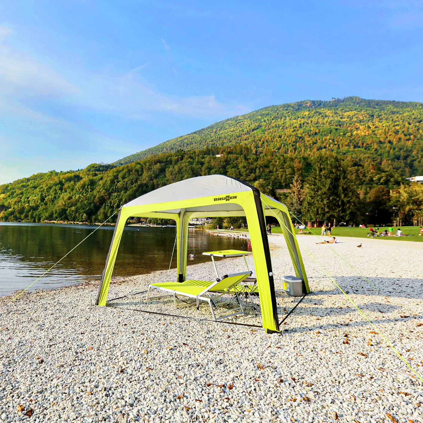 Pavillon Aquamar 300 × 220 × 300 cm, gelb-grüne Stoffstruktur mit weißem Dach, Brunner-Logo sichtbar, steht auf Kiesstrand am See; Tisch und Liege darunter