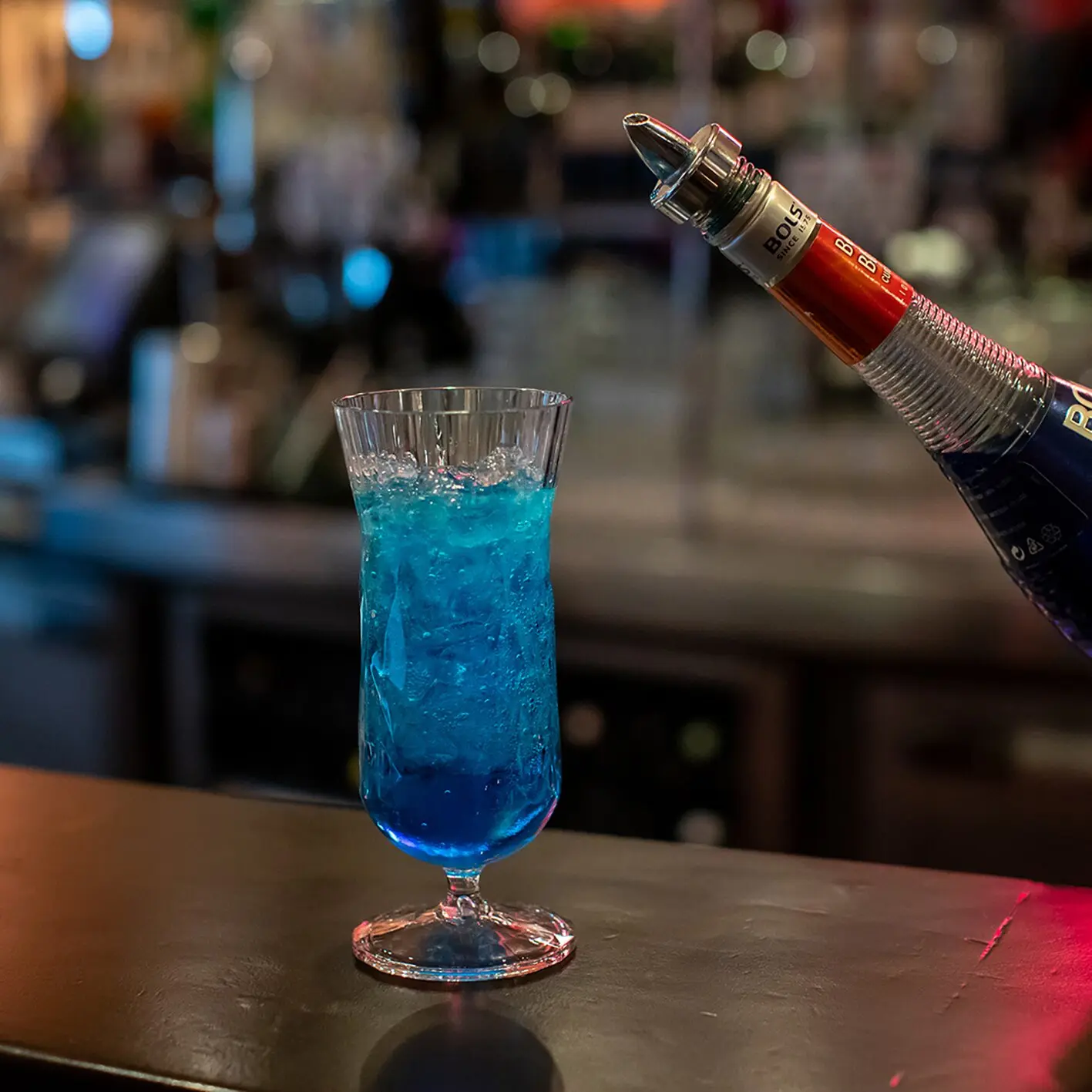 Cocktailglas mit blauem Getränk und Eis, unscharfer Barhintergrund, rechts fließt aus einer Flasche mit Ausgießer Likör ins Glas
