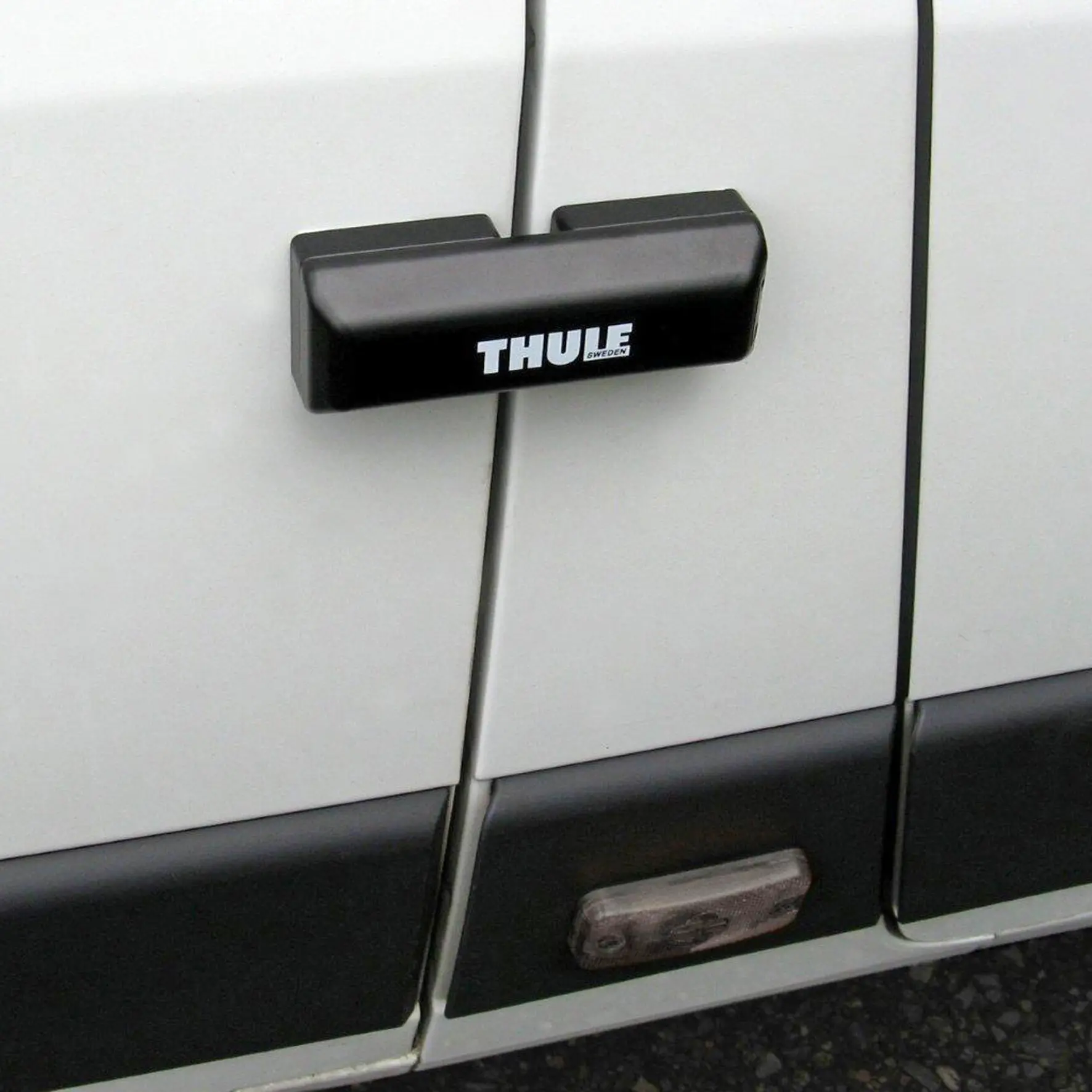 Schwarze, rechteckige Türverriegelung mit THULE-Logo, montiert an der Außenwand eines weißen Vans, sichtbar zwischen den Türblechen über der schwarzen Stoßleiste