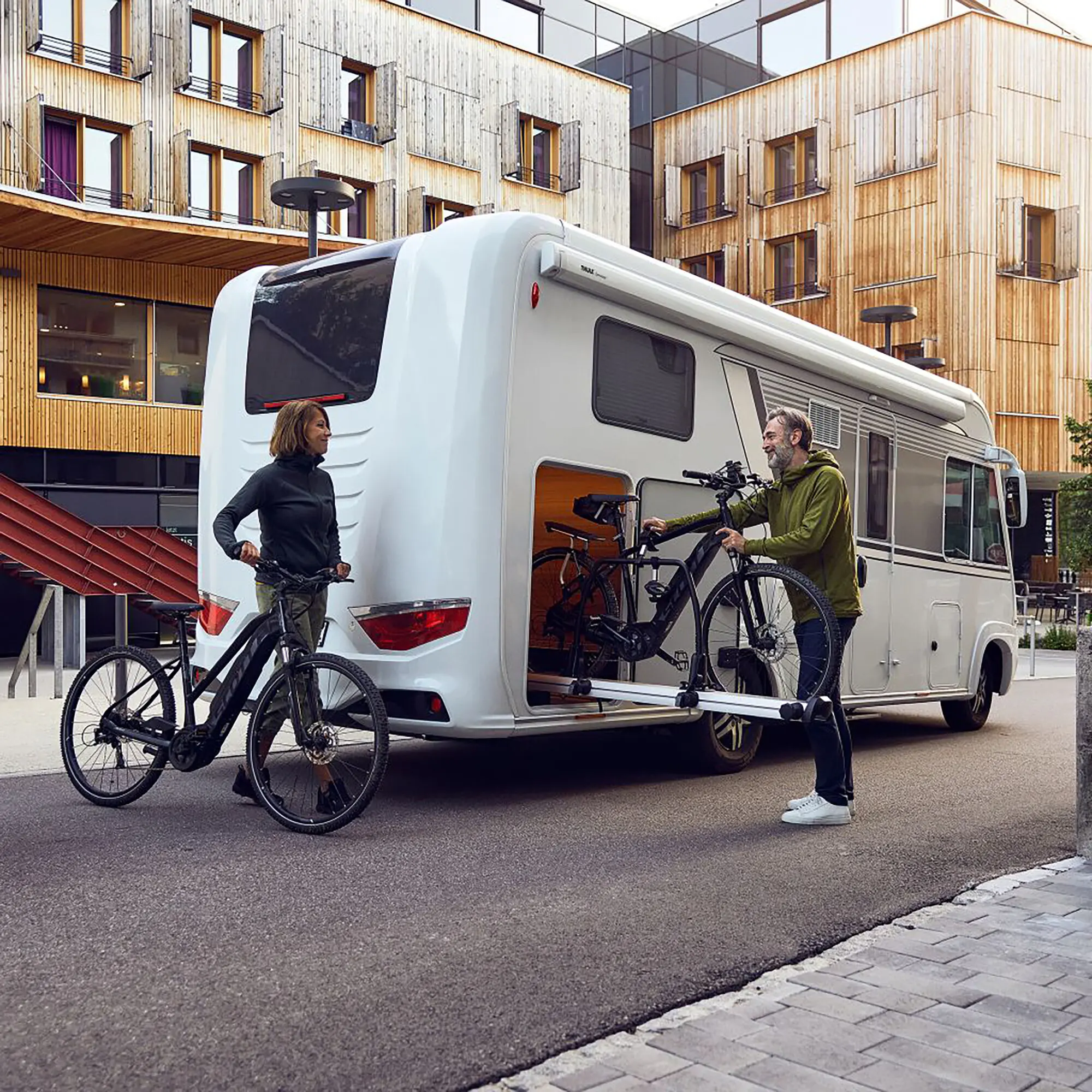 Wohnmobil mit am Heck montiertem Thule VeloSlide Short Version Fahrradheckträger, zwei Fahrräder befestigt, zwei Personen daneben