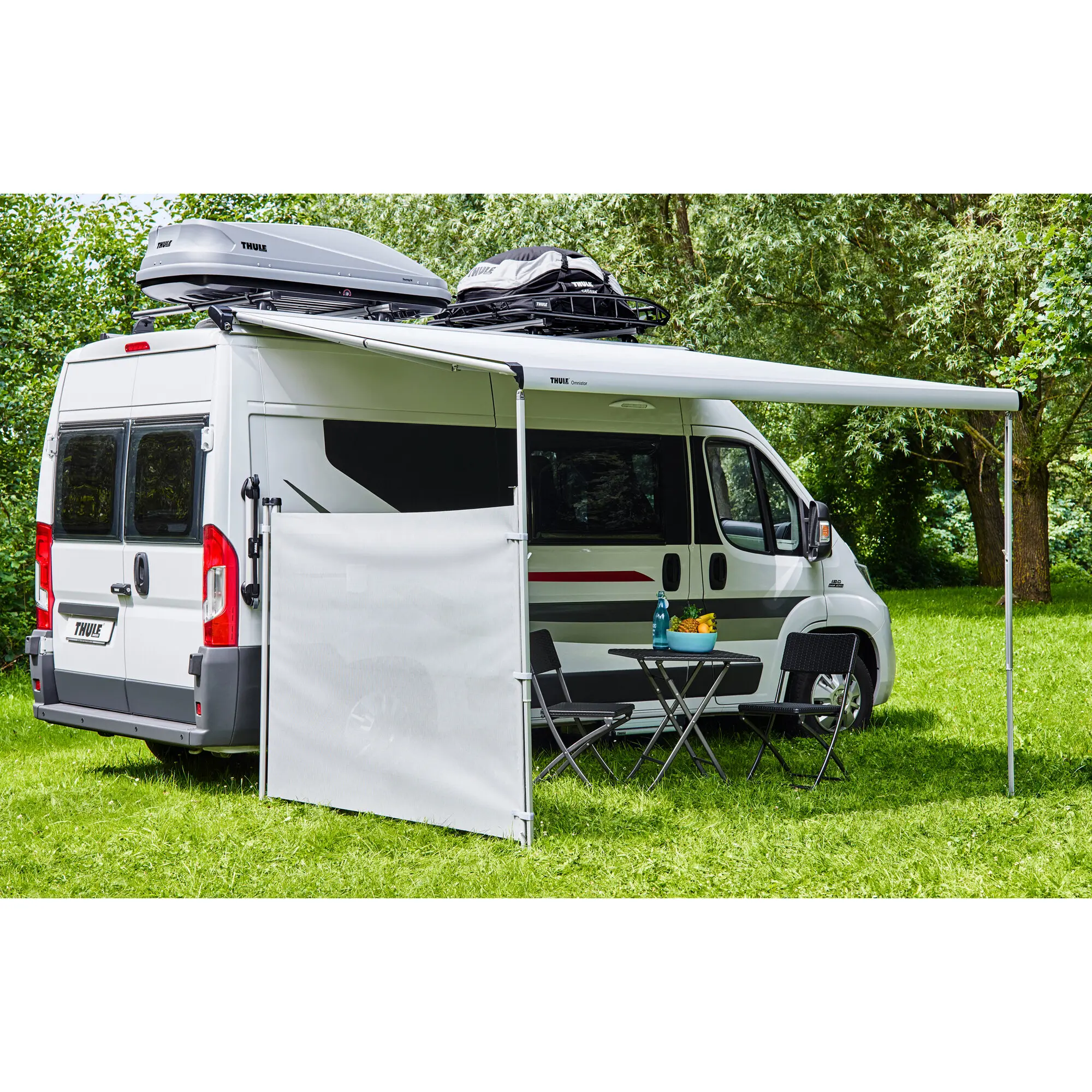 Ein weißer Campervan mit seitlichem Windschutz, davor ein Tisch und zwei Klappstühle; auf dem Dachträger graue Dachbox und schwarze Tasche