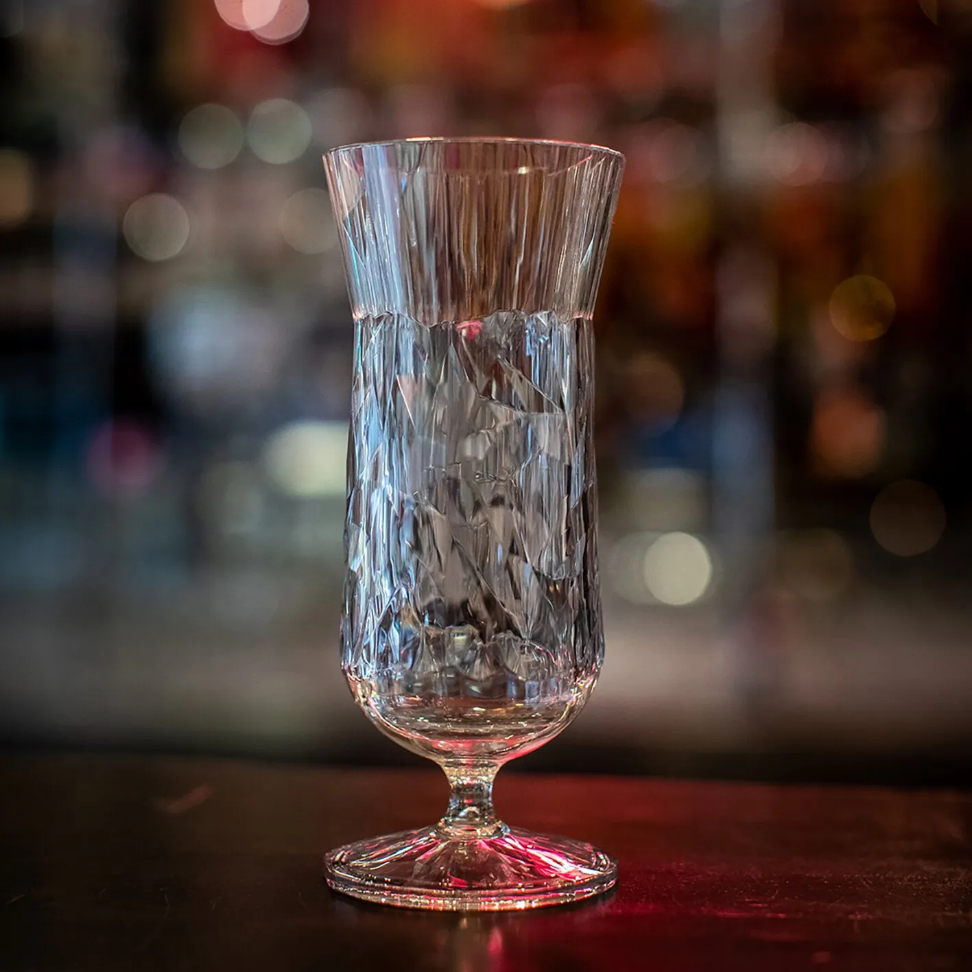 Transparentes, hohes Cocktailglas Superglas CLUB mit facettiertem Muster, kurzem Stiel und rundem Fuß auf einer Baroberfläche