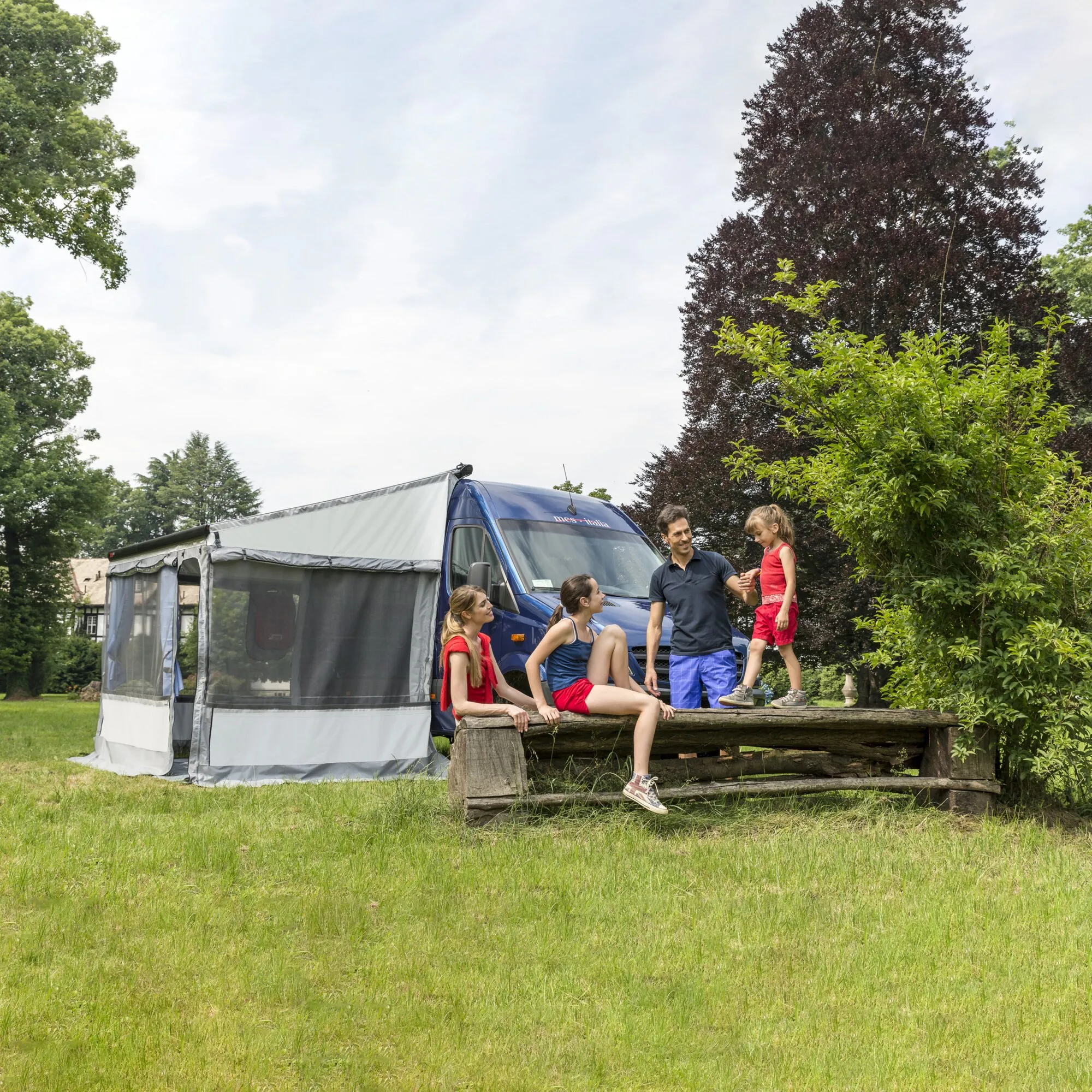 Graues Markisenzelt Privacy Room F80 an blauem Ducato H3, daneben eine Familie auf einer Wiese im Grünen