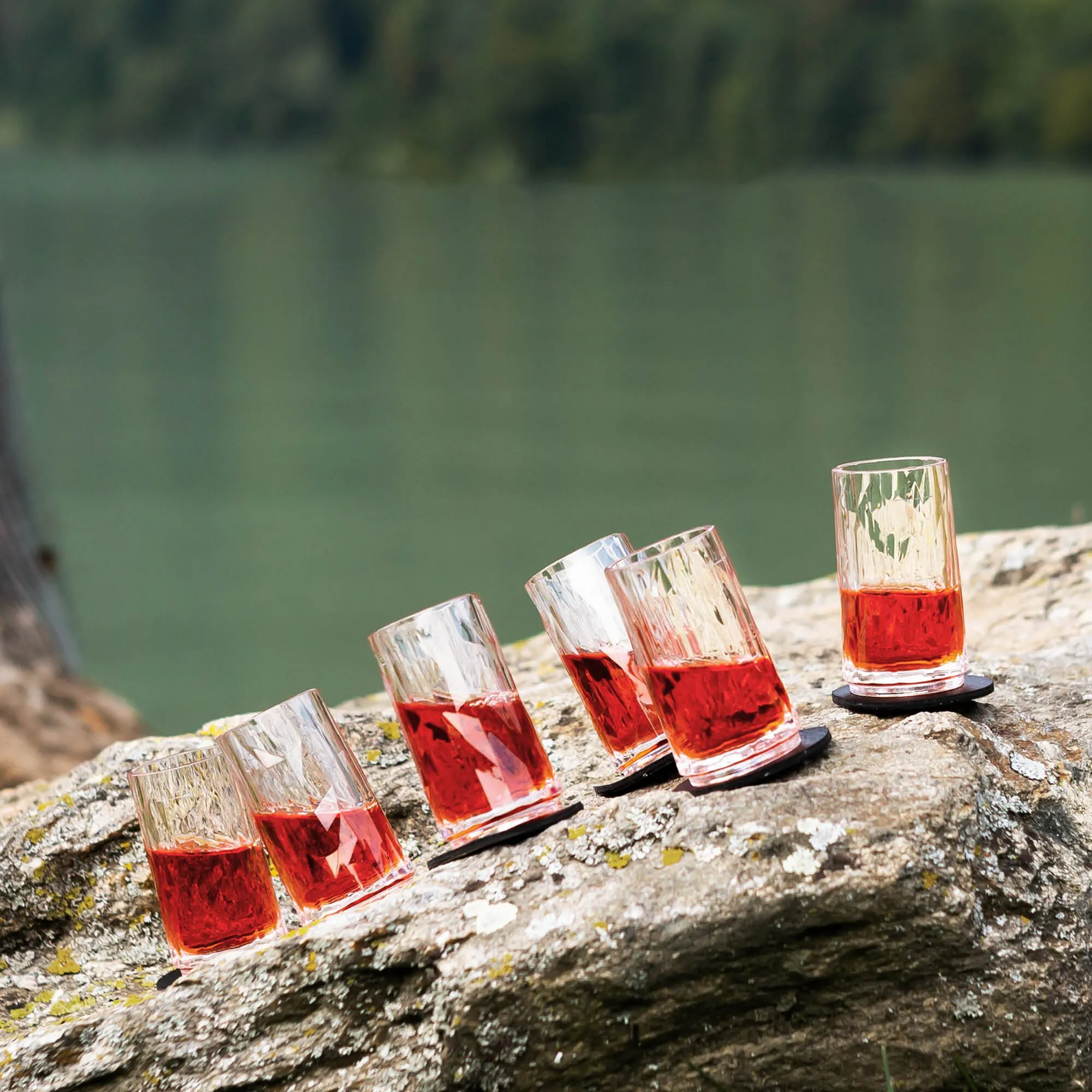 Fünf transparente Kunststoff-Schnapsgläser mit Facettenstruktur und rotem Getränk auf schwarzen Magnetunterlagen auf einem Felsen am Wasser