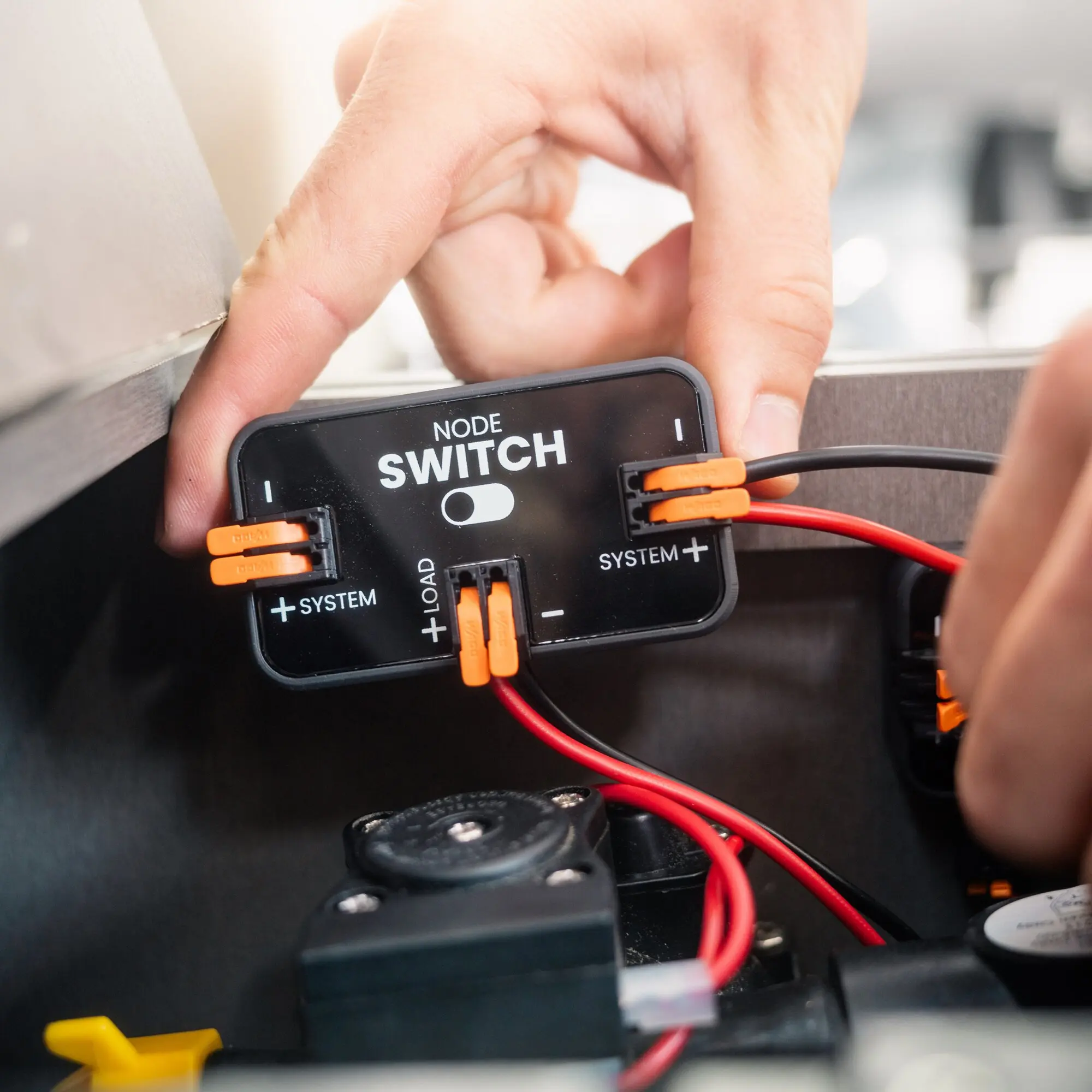 Nahaufnahme eines NODE Switch digitalen Schalters mit schwarzem Gehäuse, orange Steckkontakte und roten Kabeln, eine Hand bedient das Modul