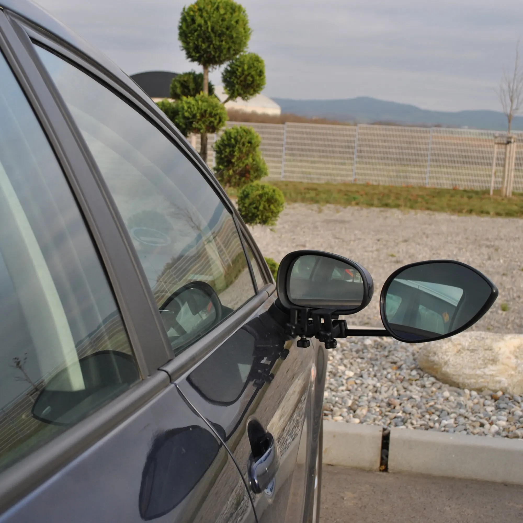 Seitenspiegel eines Autos mit einem zusätzlichen Spiegel am unteren Rand, Montagesystem sichtbar, Hintergrund Zaun und Kiesweg