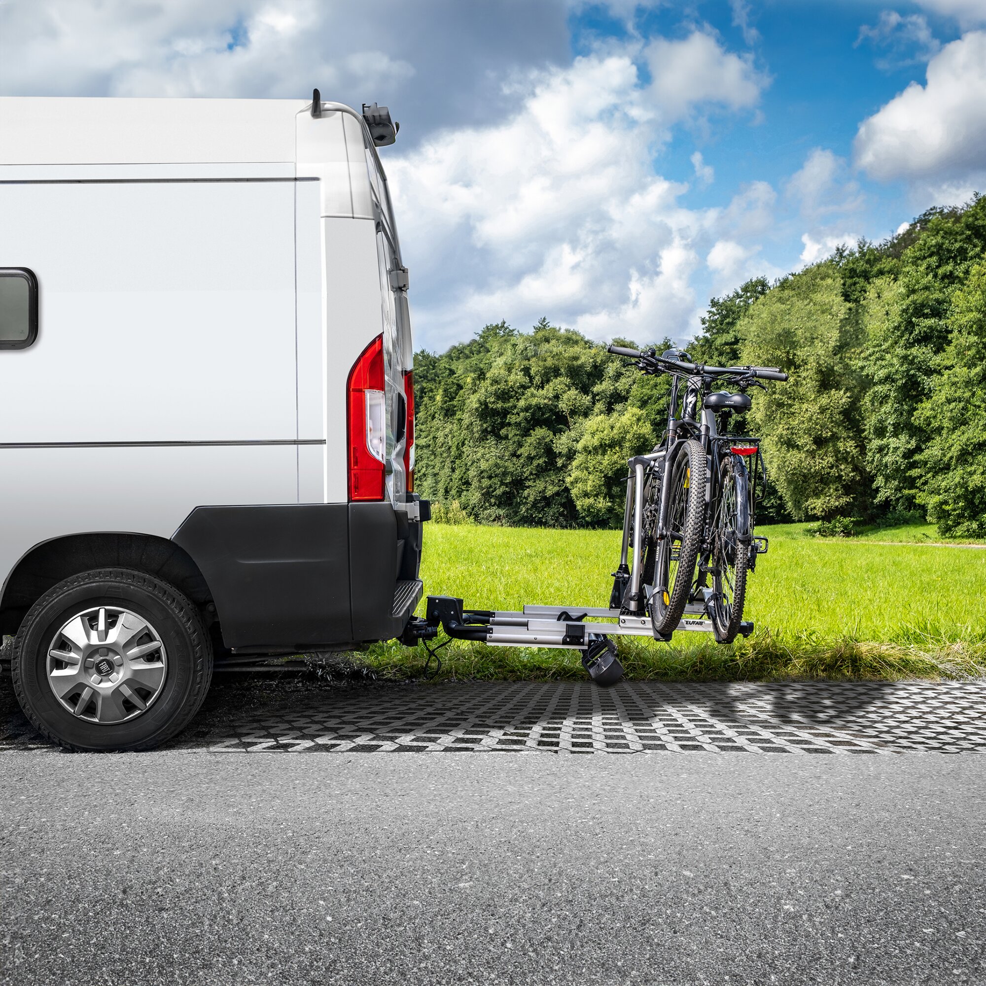 Rückseite eines weißen Transporters mit silbernem Fahrradträger an der Anhängerkupplung, zwei Mountainbikes auf dem Rack, Hintergrund grüne Wiese und Wald