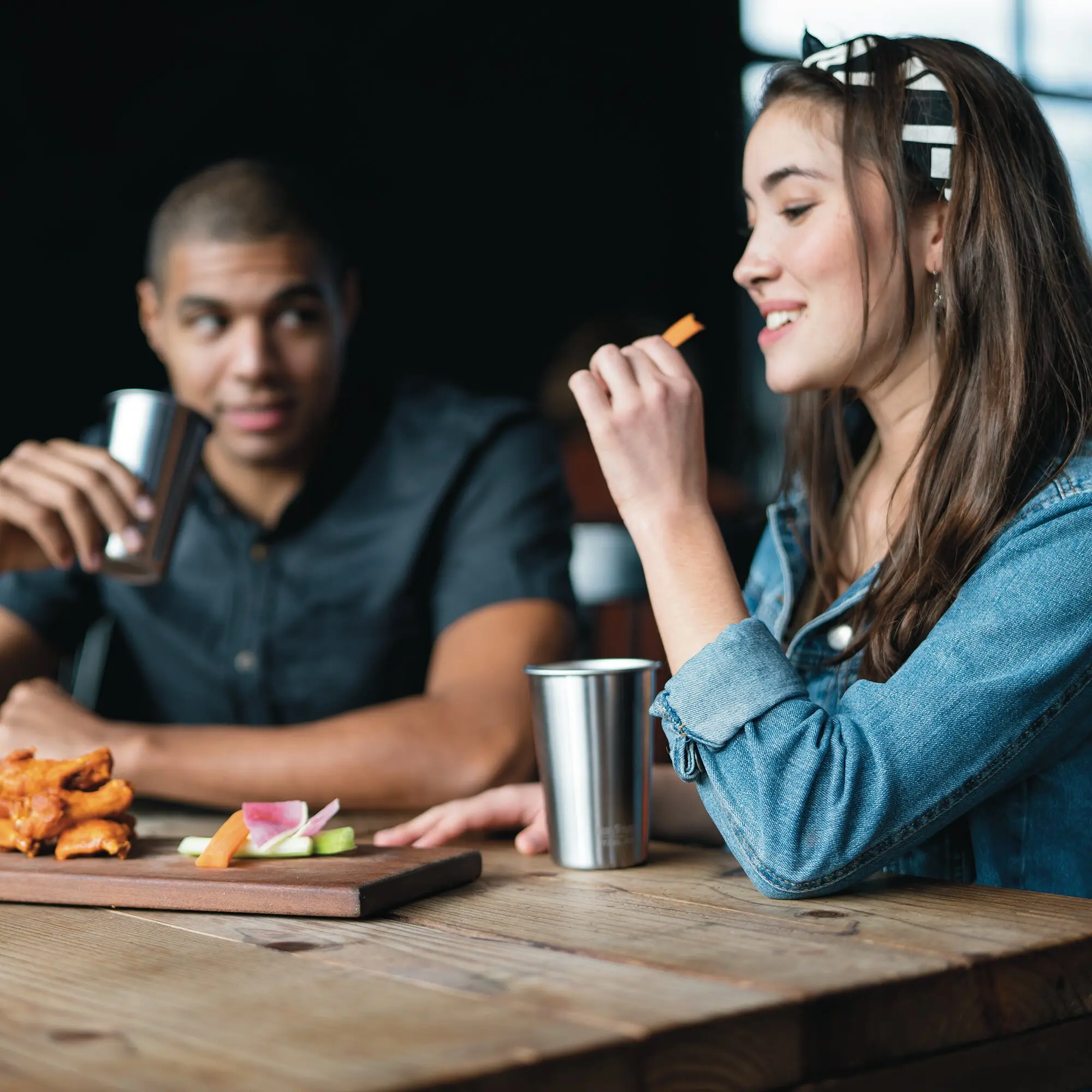 Zwei Personen sitzen an einem Holztisch und trinken aus silbernen Pint-Cups aus Edelstahl; im Vordergrund liegen Gemüse-Sticks und Snacks