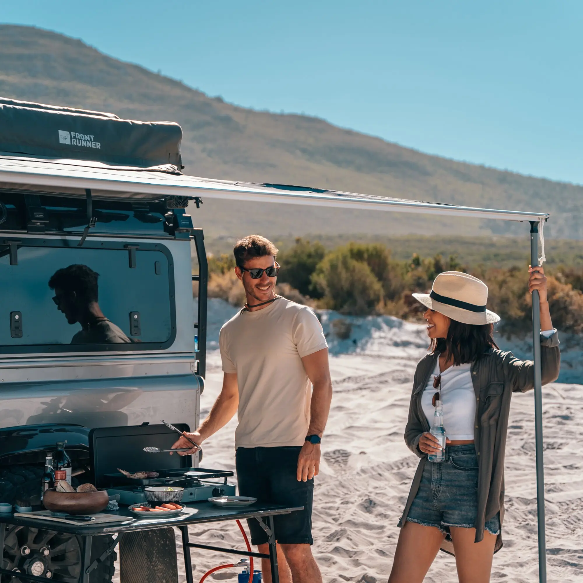 Outdoor-Küche am Heck eines Campervans, ausklappbarer Gaskocher, Teller mit Essen, zwei Reisende lachen bei Sonnenschein, Front Runner Markise im Hintergrund