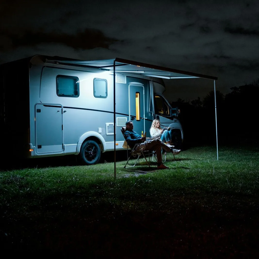 Wohnmobil bei Nacht mit ausgefahrener Markise und beleuchteter Spannstange Rafter LED, davor zwei Menschen auf Campingstühlen grasendort sitzend