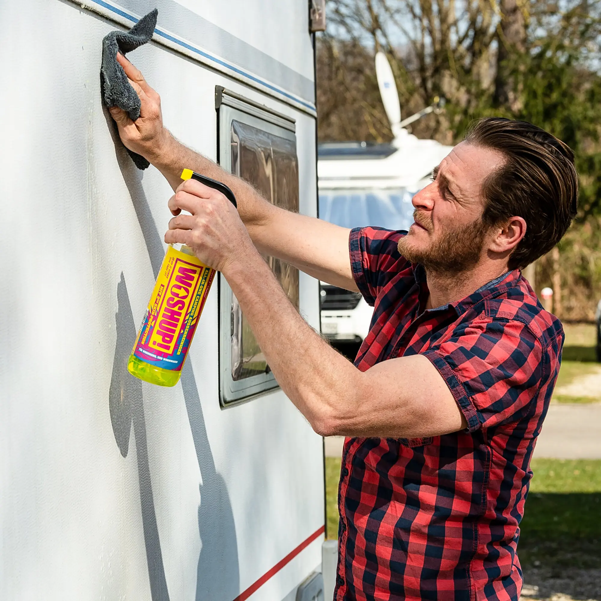 Mann reinigt die Außenwand eines Wohnwagens mit grauem Schwamm, hält gelbe Flasche Caravan Spezialreiniger in der Hand
