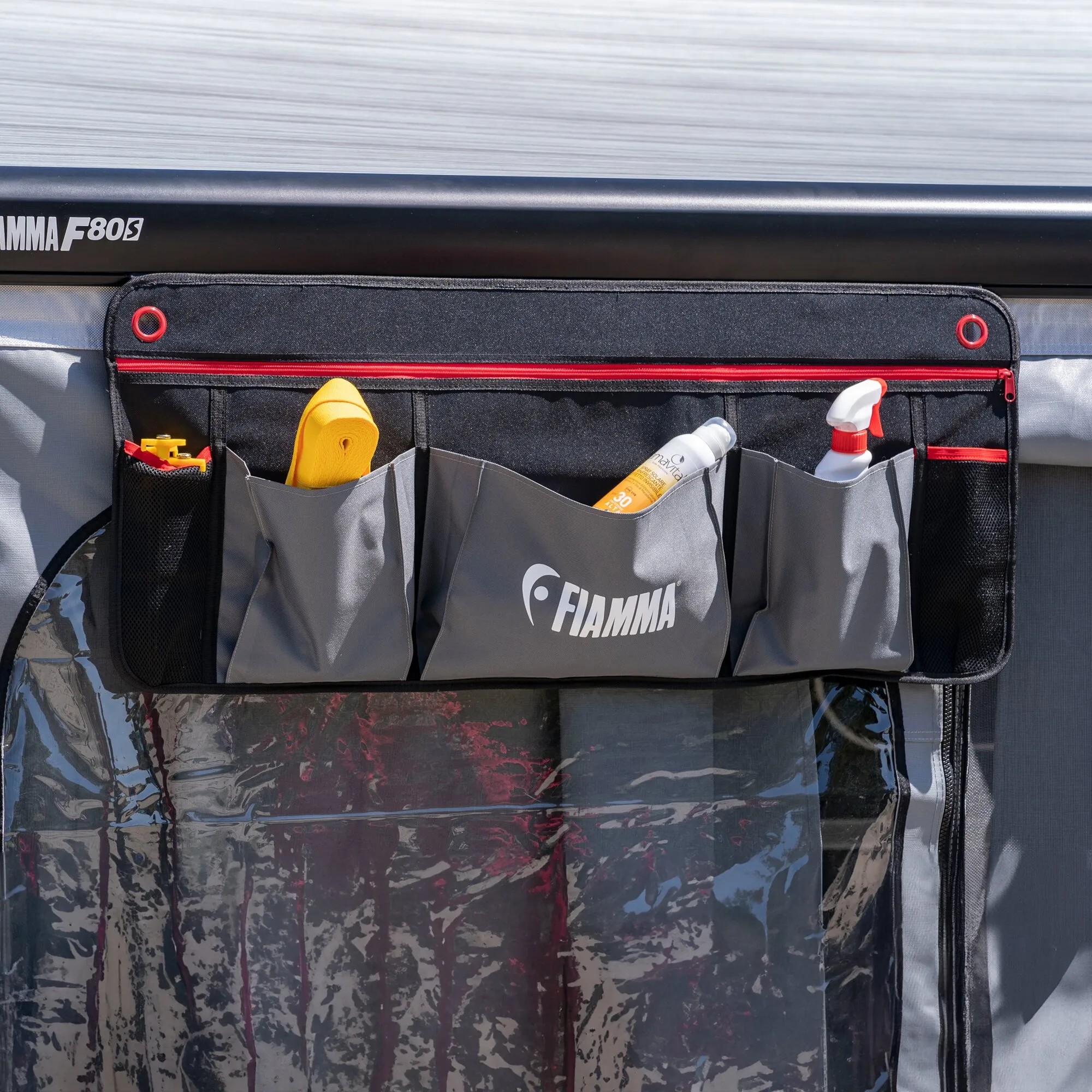 Schwarzer Fiamma Organizer L mit roten Reißverschlüssen und mehreren Fächern, gefüllt mit Sonnencreme, Sprühflasche und Zubehör am Wohnmobil