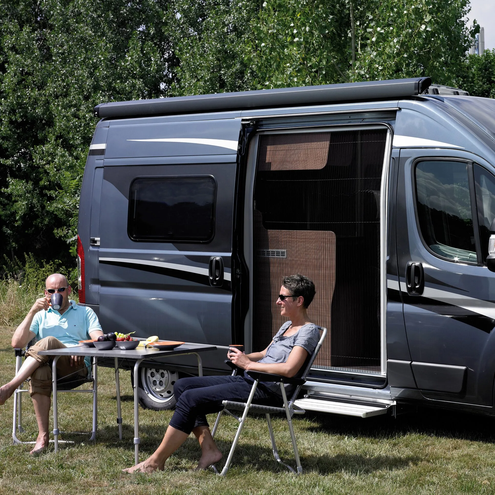 Grauer Camper mit geöffneter Schiebetür und schwarzem FlyTec FT 200 Insektenschutzgitter, davor zwei Personen beim Camping、】【
