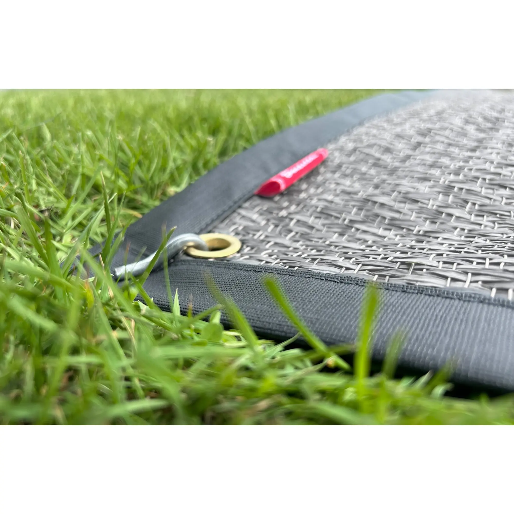 Nahaufnahme eines grauen, gewebten Outdoor-Teppichs mit Metall-Ösen; rotes Etikett liegt an der Kante, Gras im Hintergrund