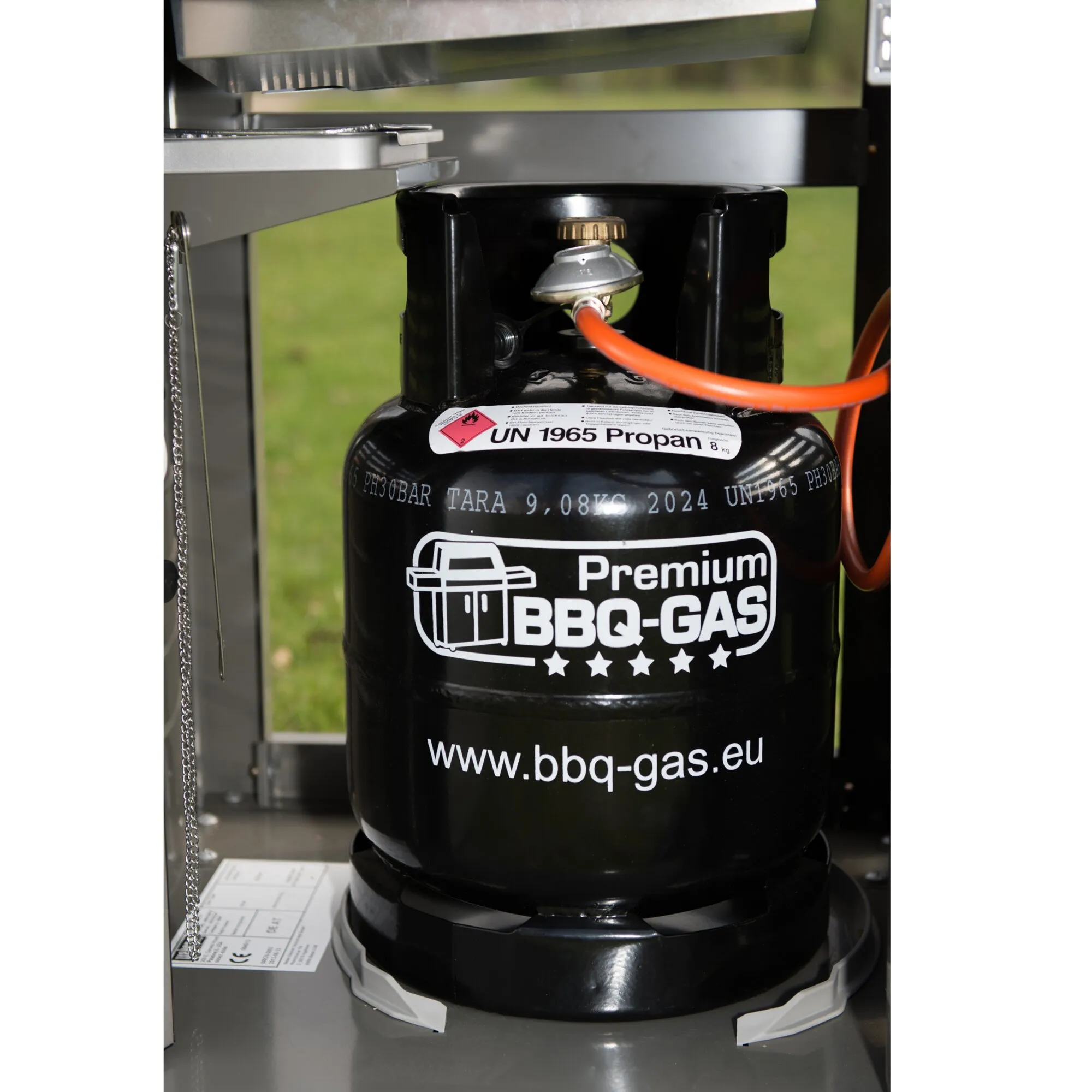 Schwarze BBQ-Gasflasche mit Druckregler und orangefarbenem Schlauch neben einem Gasgrill, beschriftet mit UN 1965 Propan und 8 kg