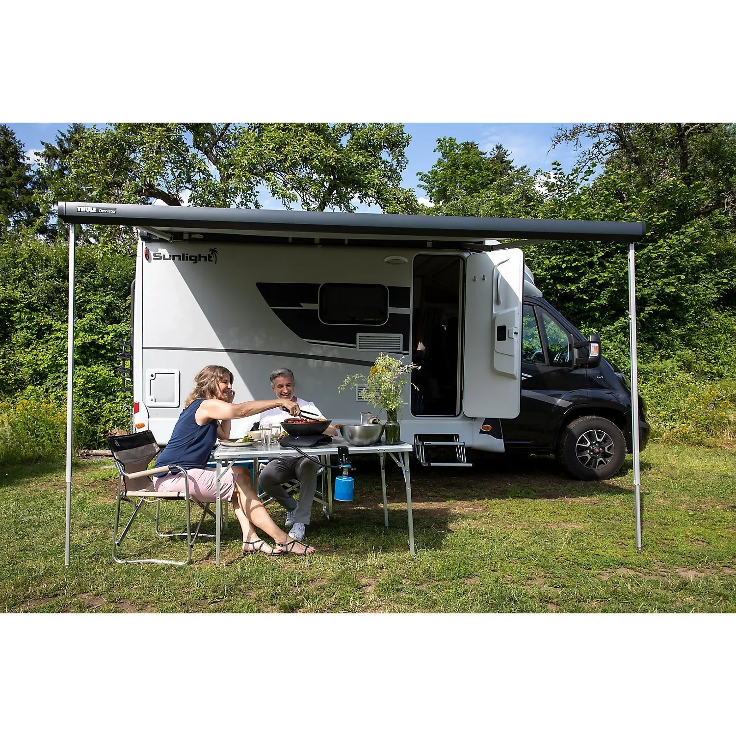 Tischgrill 360 Grill CV mit Grillgut auf einem Campingtisch vor einem Wohnmobil, während ein Paar gemeinsam grillt