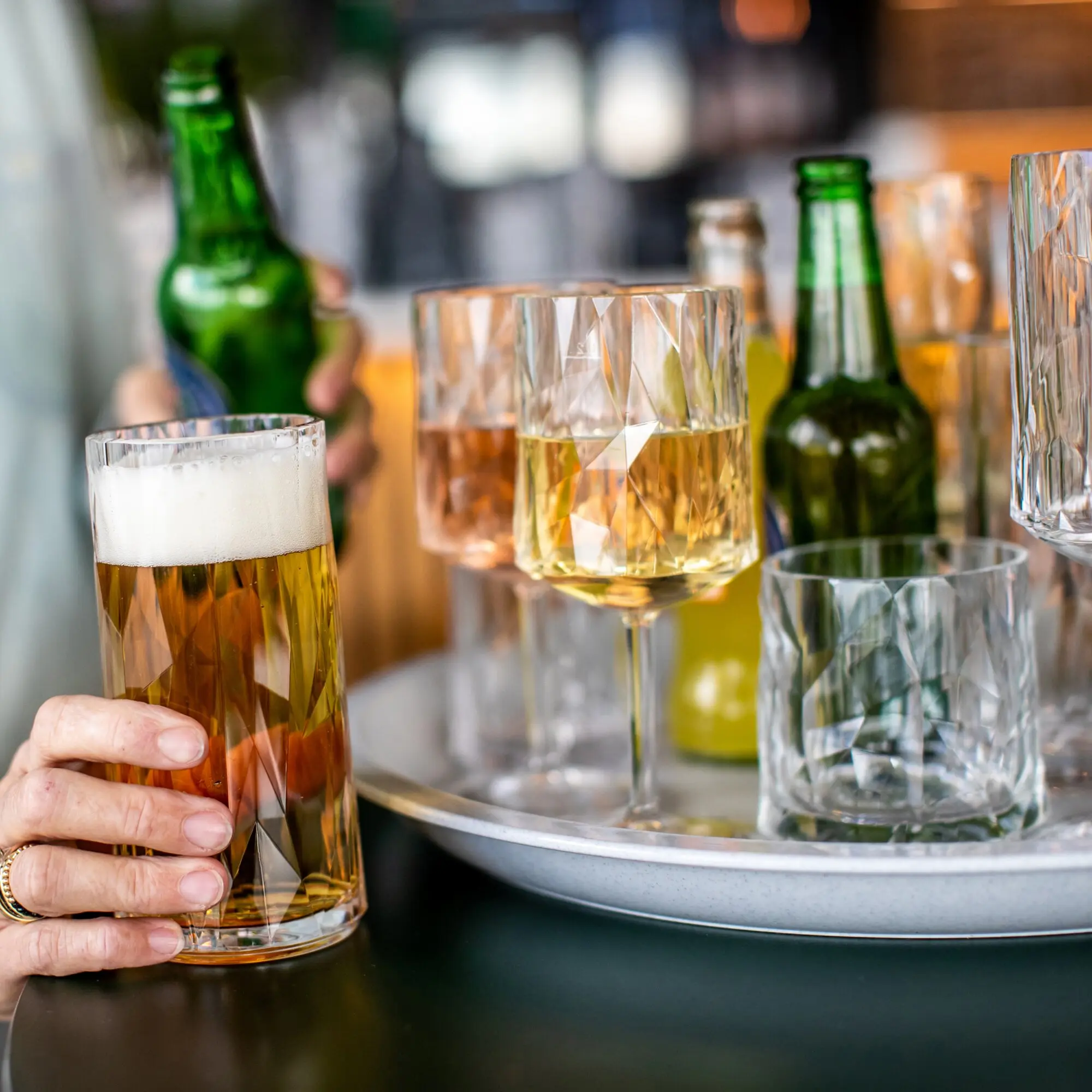 Nahaufnahme eines facettierten Superglas CLUB Longdrink-Glases mit bernsteinfarbener Flüssigkeit, von einer Hand gehalten; im Hintergrund grüne Flaschen