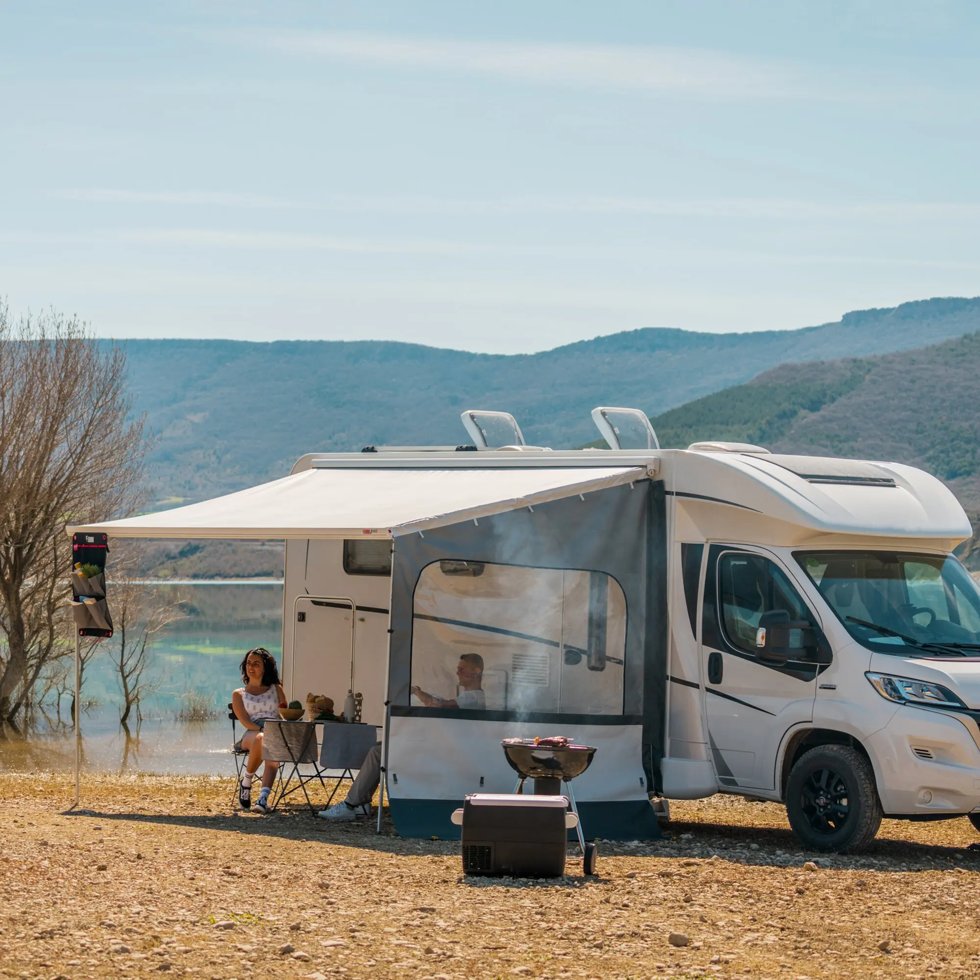 Camper mit ausgefahrener Seitenwand-Markise, davor zwei Outdoor-Sessel an einem Tisch, Frau sitzt, Mann im Innenraum sichtbar, See und Berge im Hintergrund
