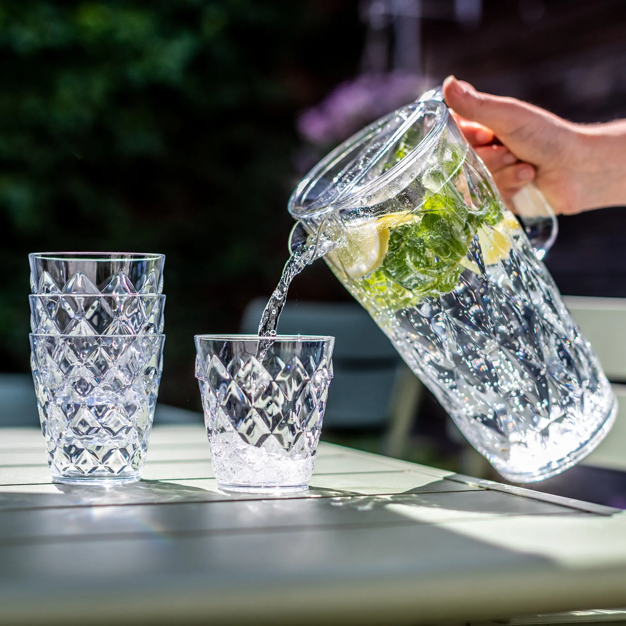 Transparente CRYSTAL-Kanne mit Zitronen und Minze schenkt Wasser in einen strukturierten Becher ein, drei weitere Becher stehen daneben
