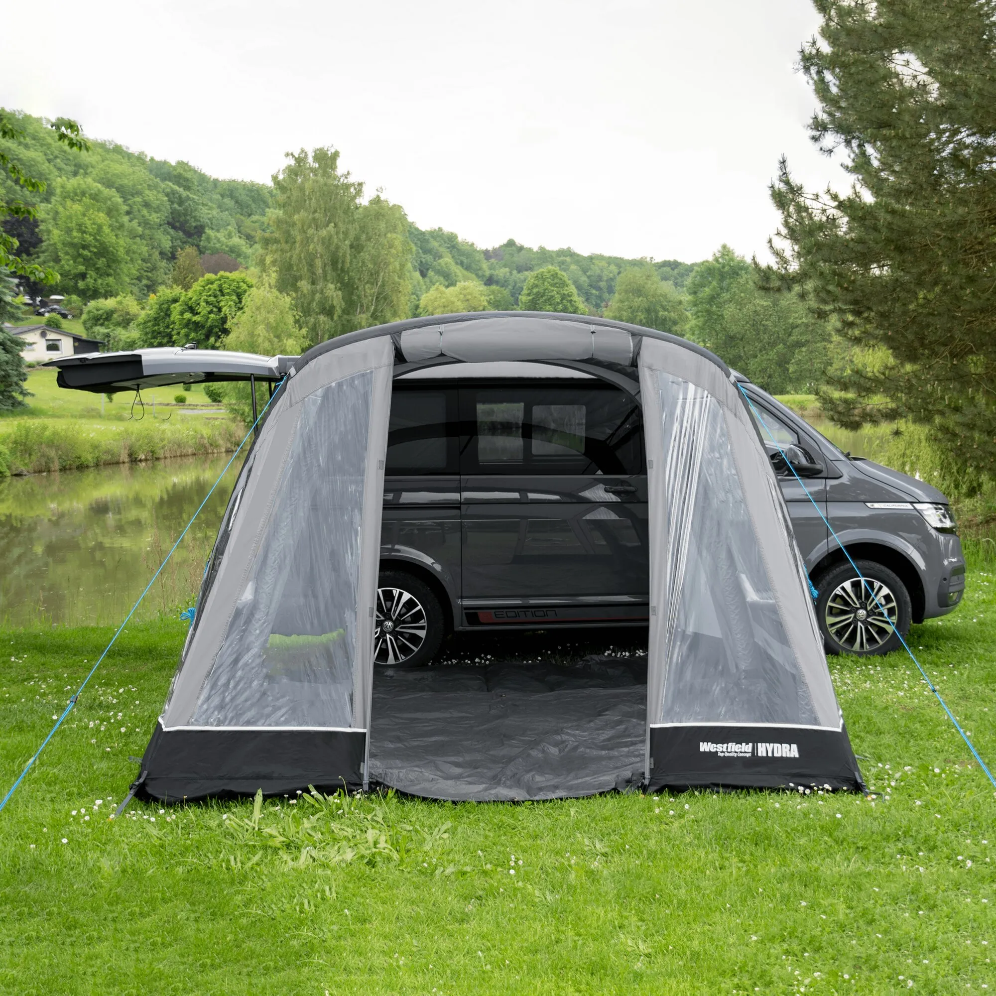 Graues Teilzelt Hydra von Westfield mit transparenter Front und Abspannleinen, an einen dunklen Volkswagen angebaut auf einer Wiese am See