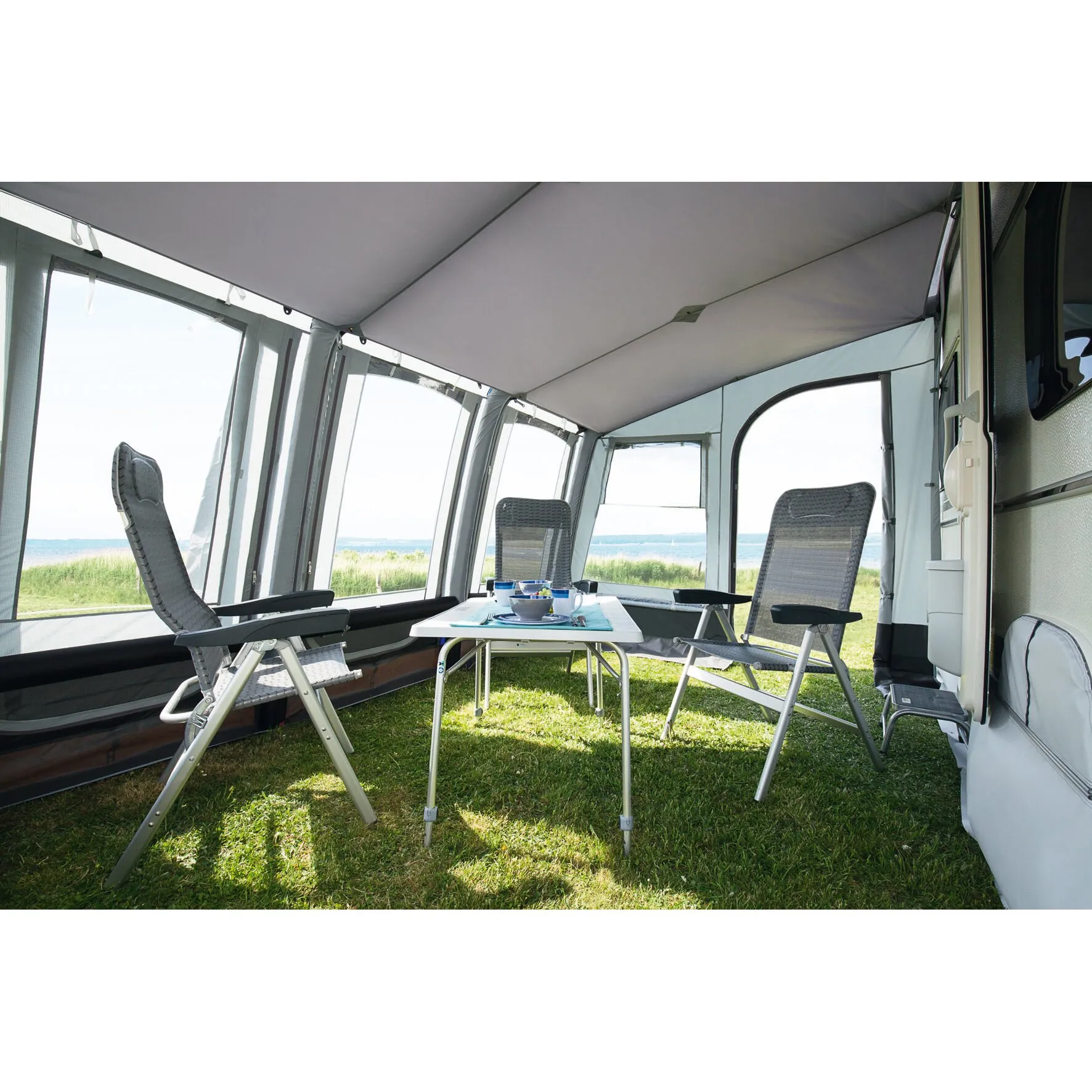 Innenansicht des Teilzelts Space Air HQ am Wohnwagen mit grauem Dach, Fenstern, drei Klappstühlen und gedecktem Tisch auf einer Wiese am Meer