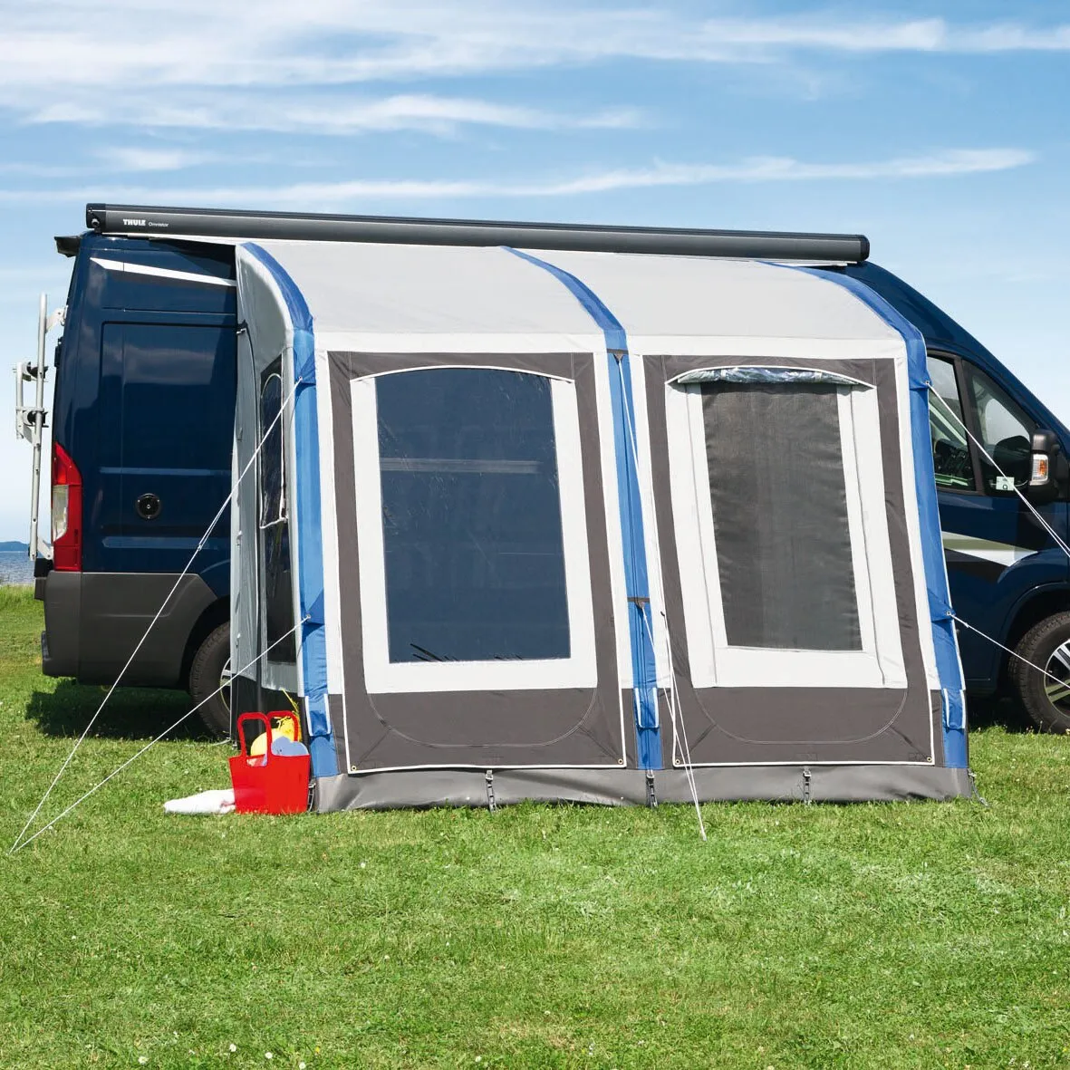 Blau-weißes Teilzelt Space Air HQ am dunkelblauen Campervan, mit zwei großen Fenstern und Abspannleinen auf einer Wiese