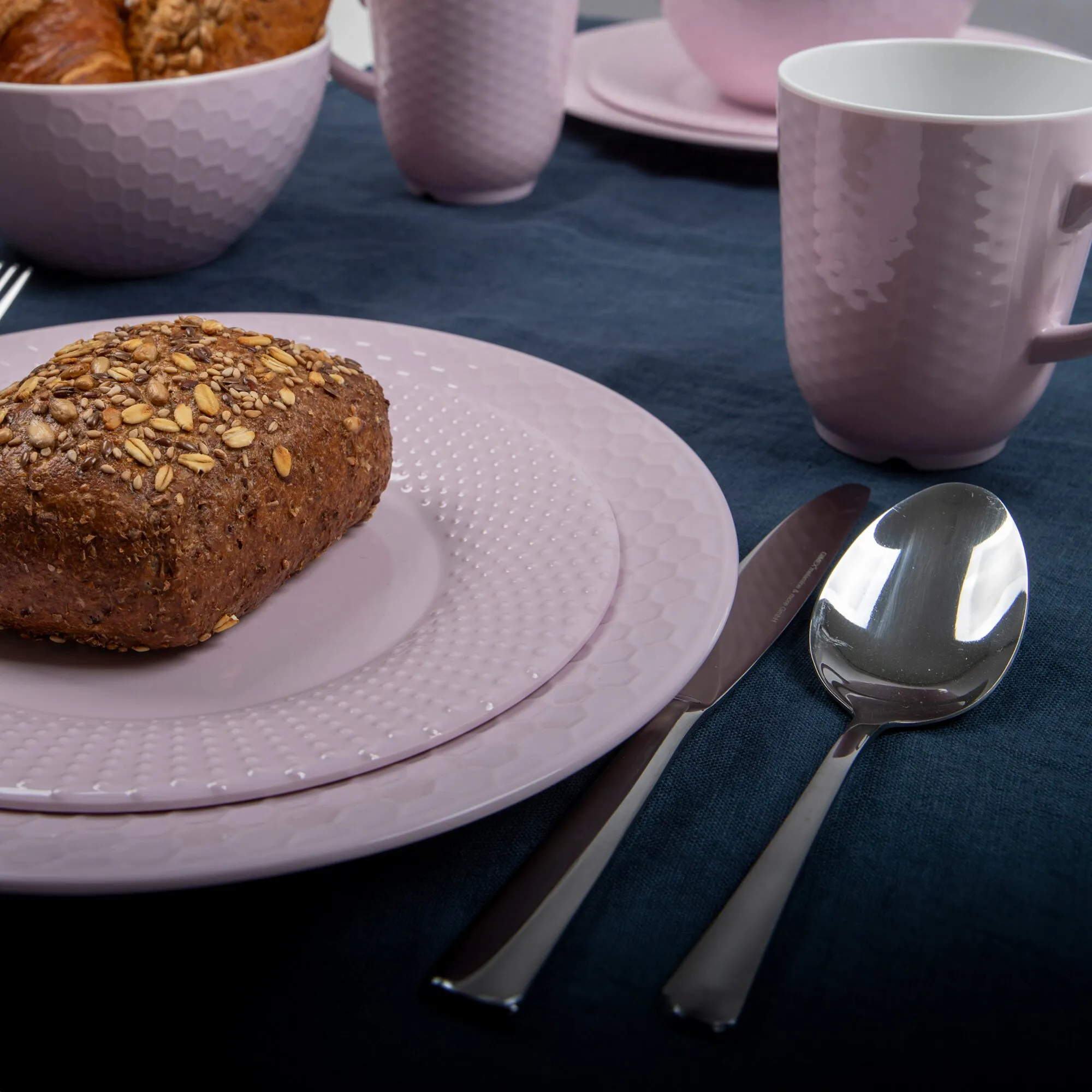Rosa Geschirrset Royal Line mit strukturierten Tellern, Schalen und Tassen, angerichtet mit Brot sowie Besteck auf blauem Tischtuch