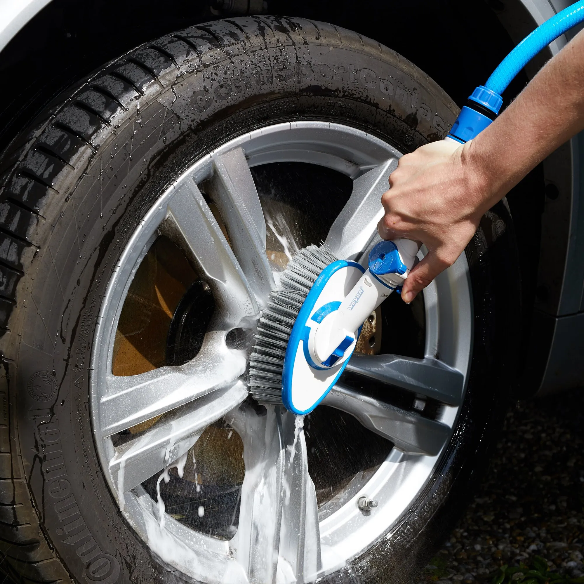 Weiße ovale Waschbürste mit blauem Rand und drehbarem Bürstenkopf reinigt einen nassen, eingeschäumten Autofelgenkranz