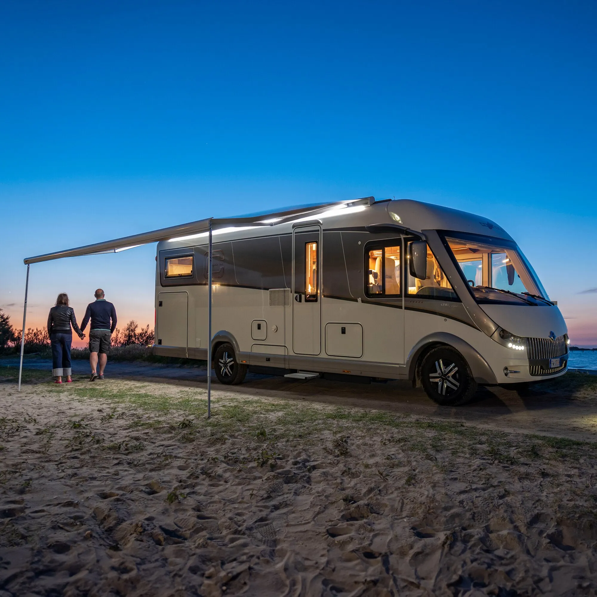 Wohnmobil mit ausgefahrener Markise und eingeschalteter Strip-Beleuchtung am Strand bei Abenddämmerung, zwei Personen im Hintergrund