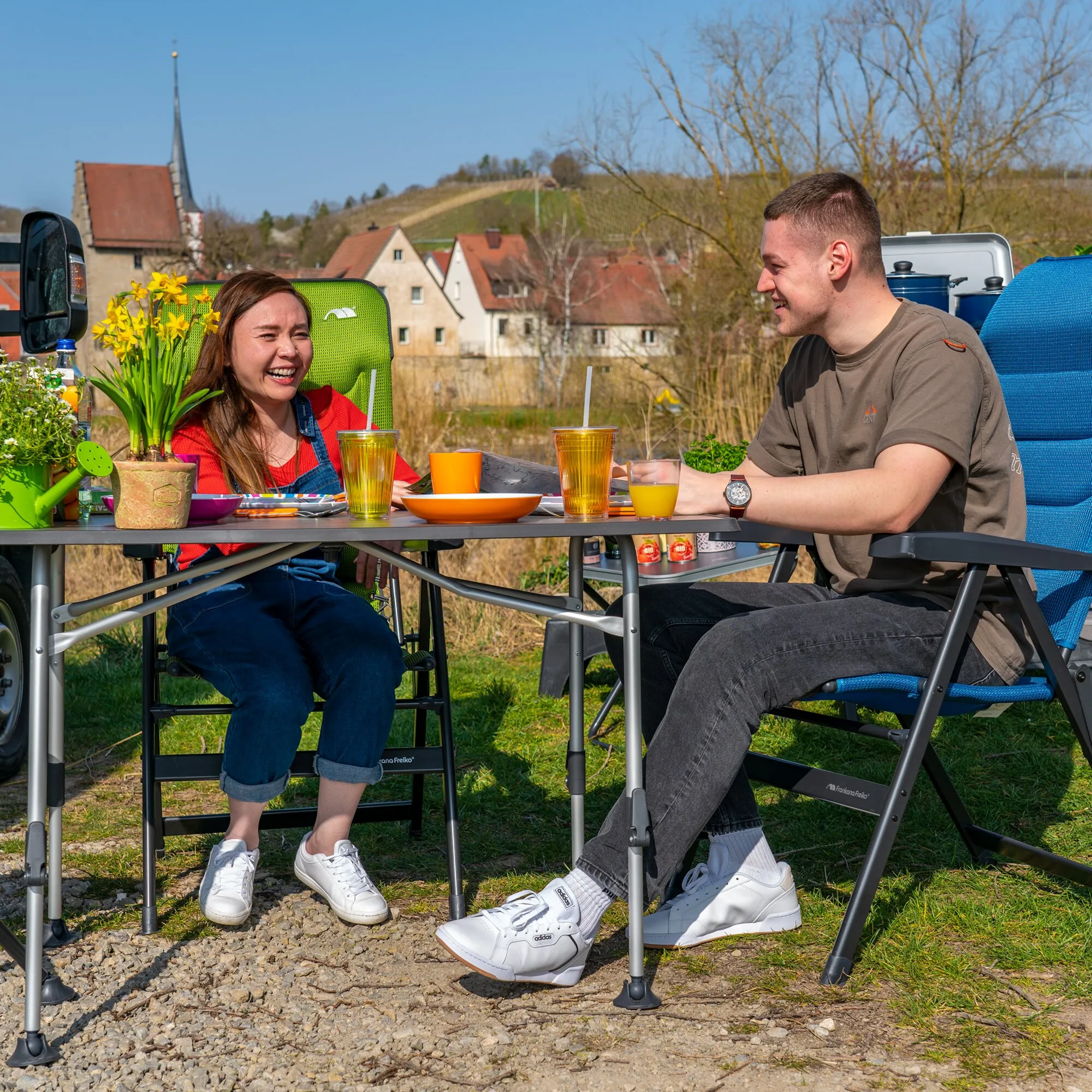 Schwarzer Campingtisch HighQ Blackline mit Getränken und Geschirr, an dem zwei Personen im Freien sitzen und essen