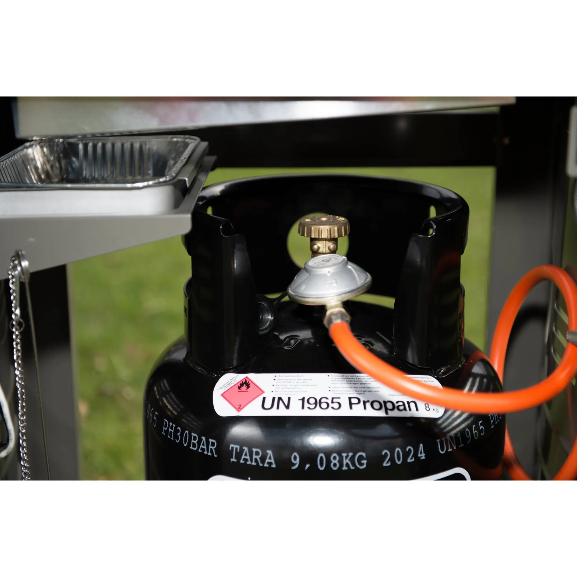 Schwarze BBQ-Gasflasche mit Druckregler, orangefarbenem Gasschlauch und sichtbarer Kennzeichnung UN 1965 Propan neben dem Grill