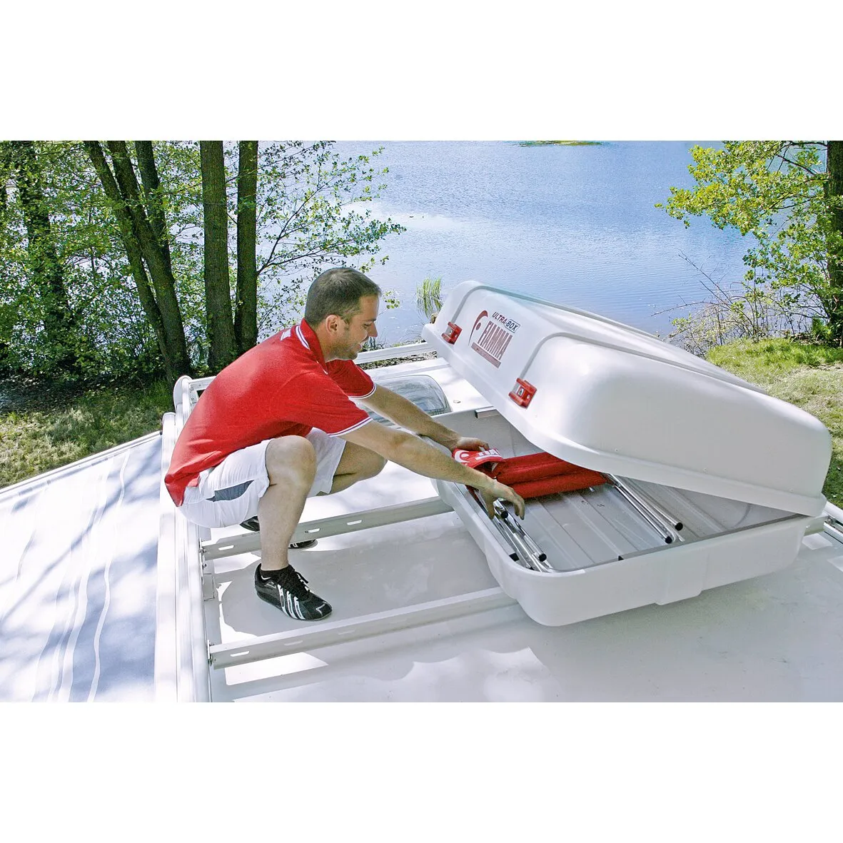 Mann befestigt eine geöffnete weiße Ultra-Box-Dachbox mit roten Verschlüssen auf einem Fahrzeugdach am See