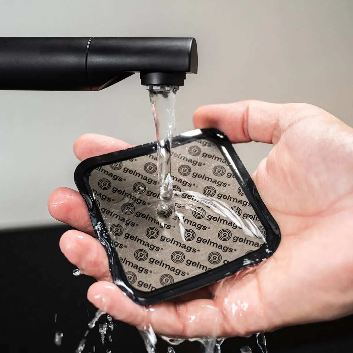 Hand hält ein schwarzes quadratisches Nano-Gel-Pads 2.0 Befestigungspad mit wiederholtem gelmags-Muster, unter fließendem Wasser aus dem Hahn