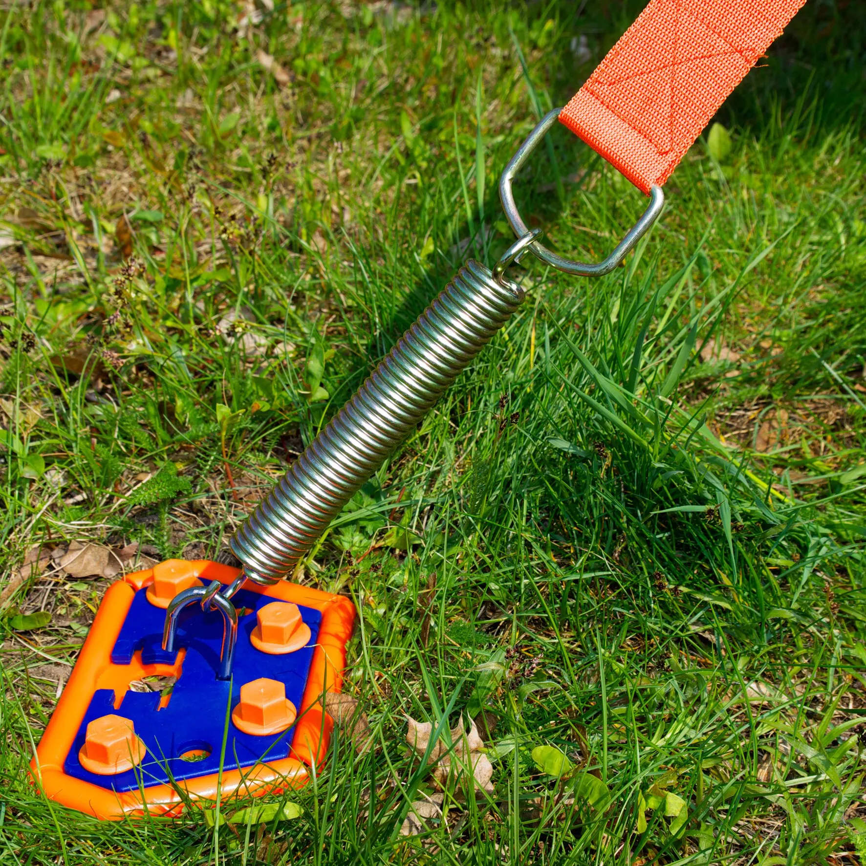 Orange-blaue PP-Ankerplatte im Gras, verbunden mit einer glänzenden Metallfeder und einem orangefarbenen Gurt