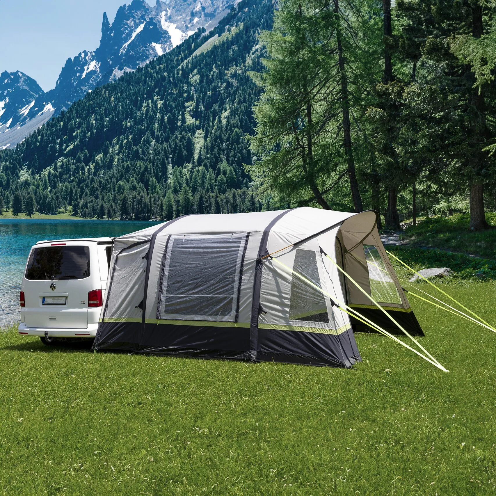 Graues Busvorzelt Advantourer an einem weißen Campingbus auf einer Wiese am Bergsee vor bewaldeten Alpenfelsgipfeln