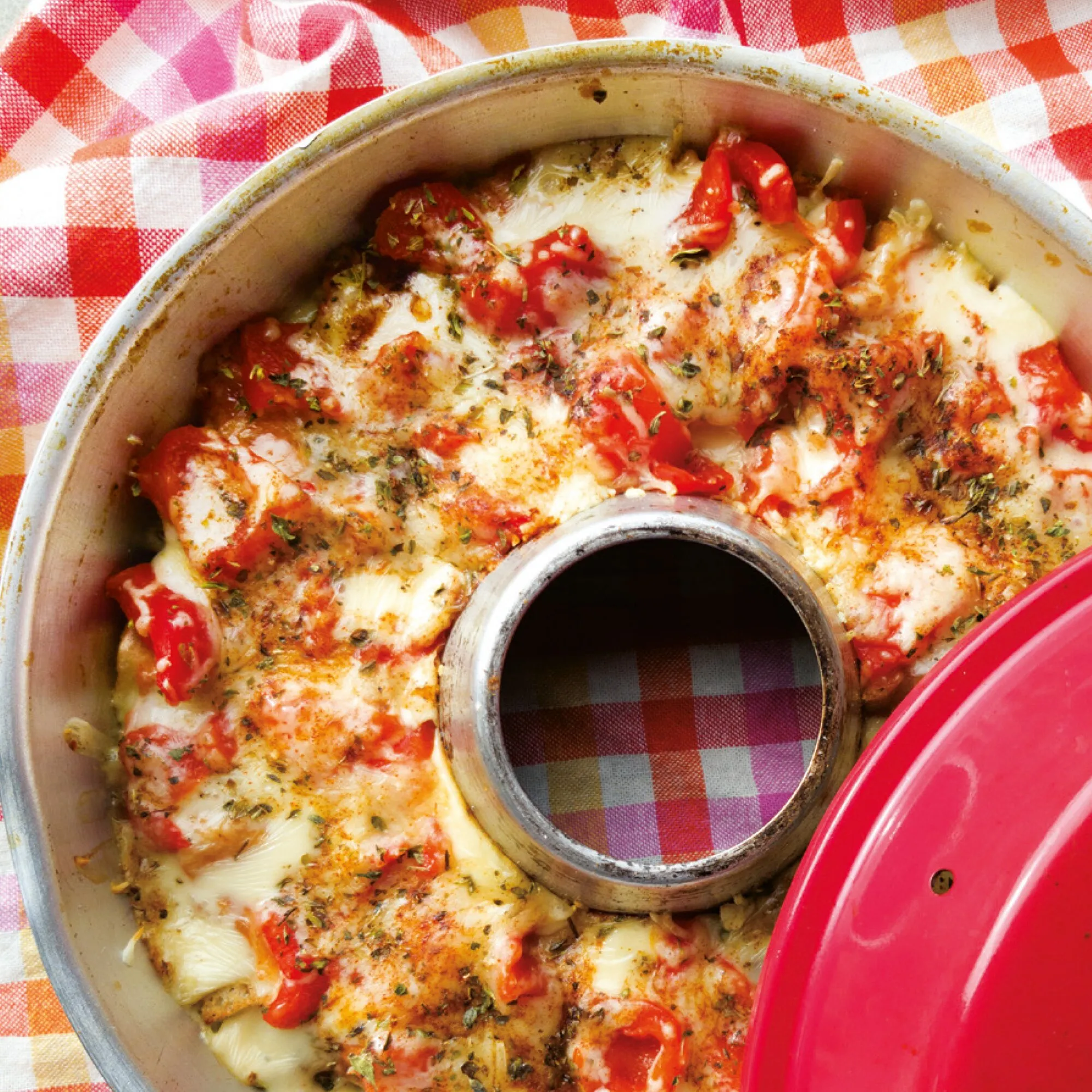 Überbackenes One-Pot-Gericht mit Tomaten, geschmolzenem Käse und Kräutern im runden Omnia-Backofen mit rotem Deckel