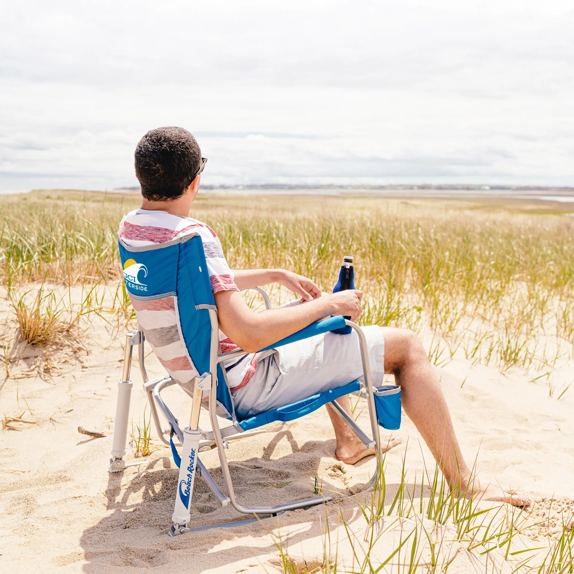 Mann sitzt in blauem Camping-Schaukelstuhl Beach Rocker™ am Strand und blickt auf das Meer