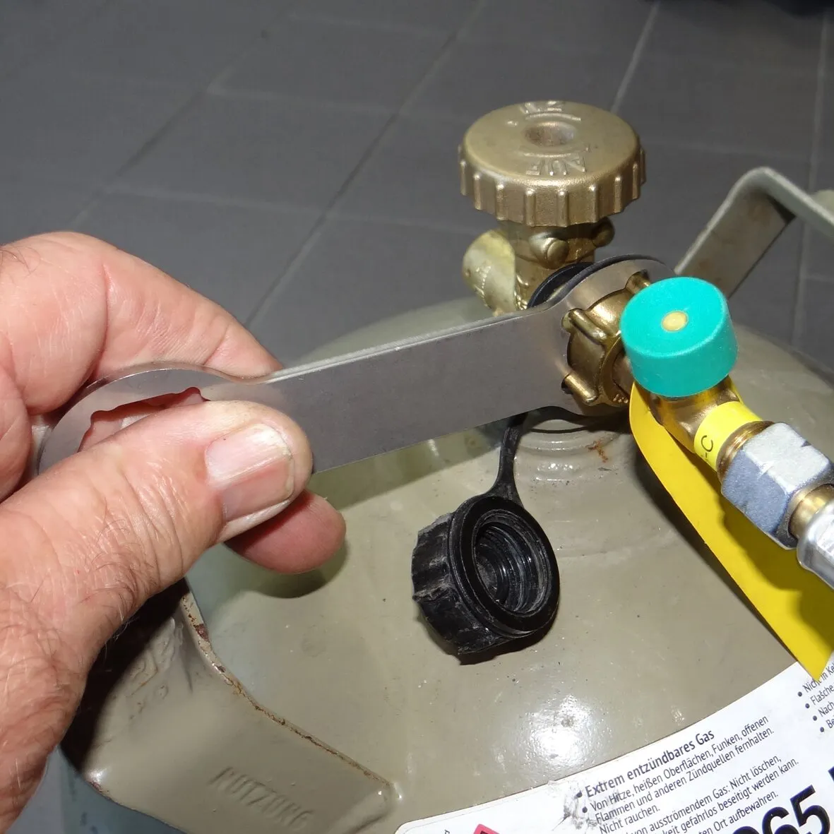 Hand gehaltener grauer Gasflaschenschlüssel beim Anziehen eines Gasreglers an einer Gasflasche mit gelbem Schlauch