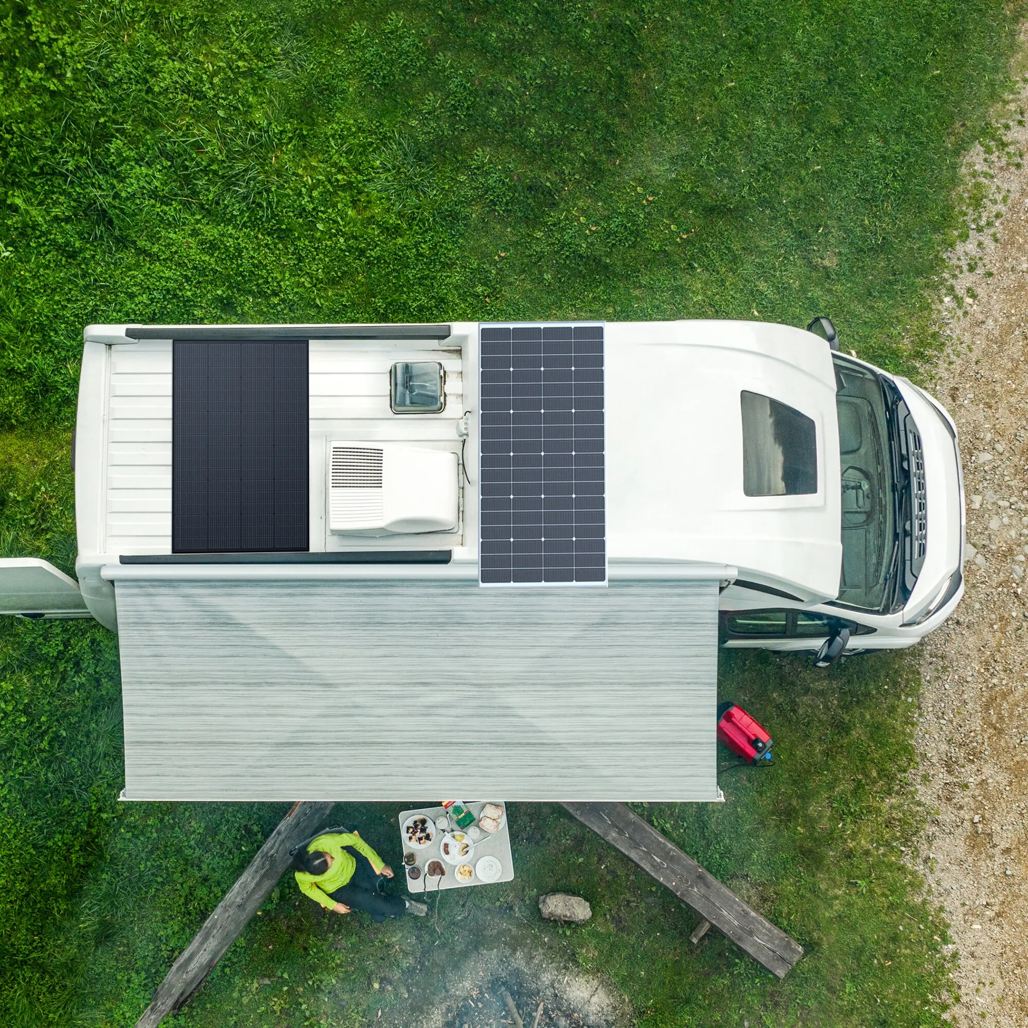 Vogelperspektive auf das Dach eines weißen Wohnmobils mit zwei Solarmodulen, Dachluke und Belüftungsgitter, umgeben von grünem Gras und einem Kiesweg