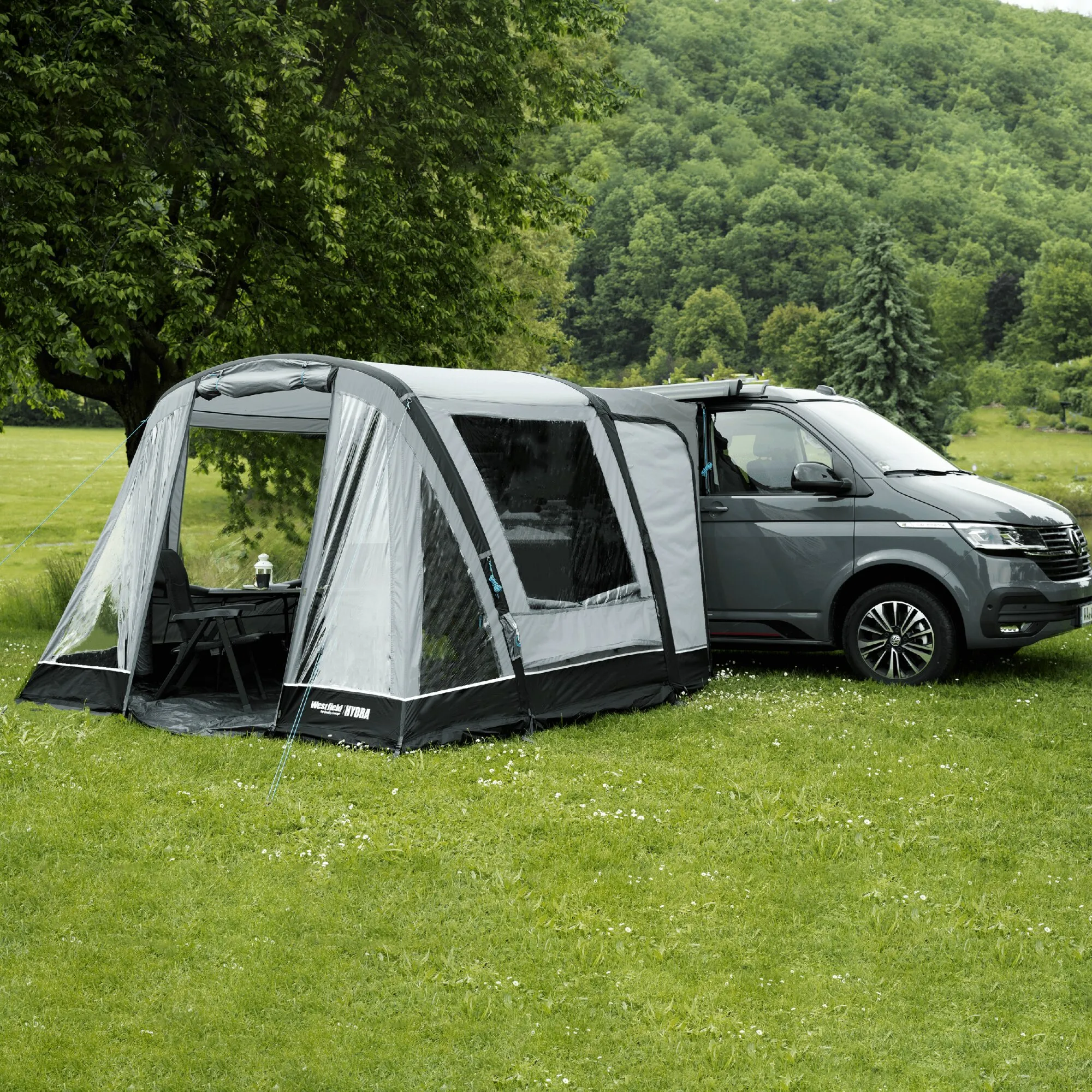 Graues Teilzelt Hydra am Heck eines dunklen Vans auf einer Wiese, mit transparenten Fenstern, Abspannleinen und Campingmöbeln
