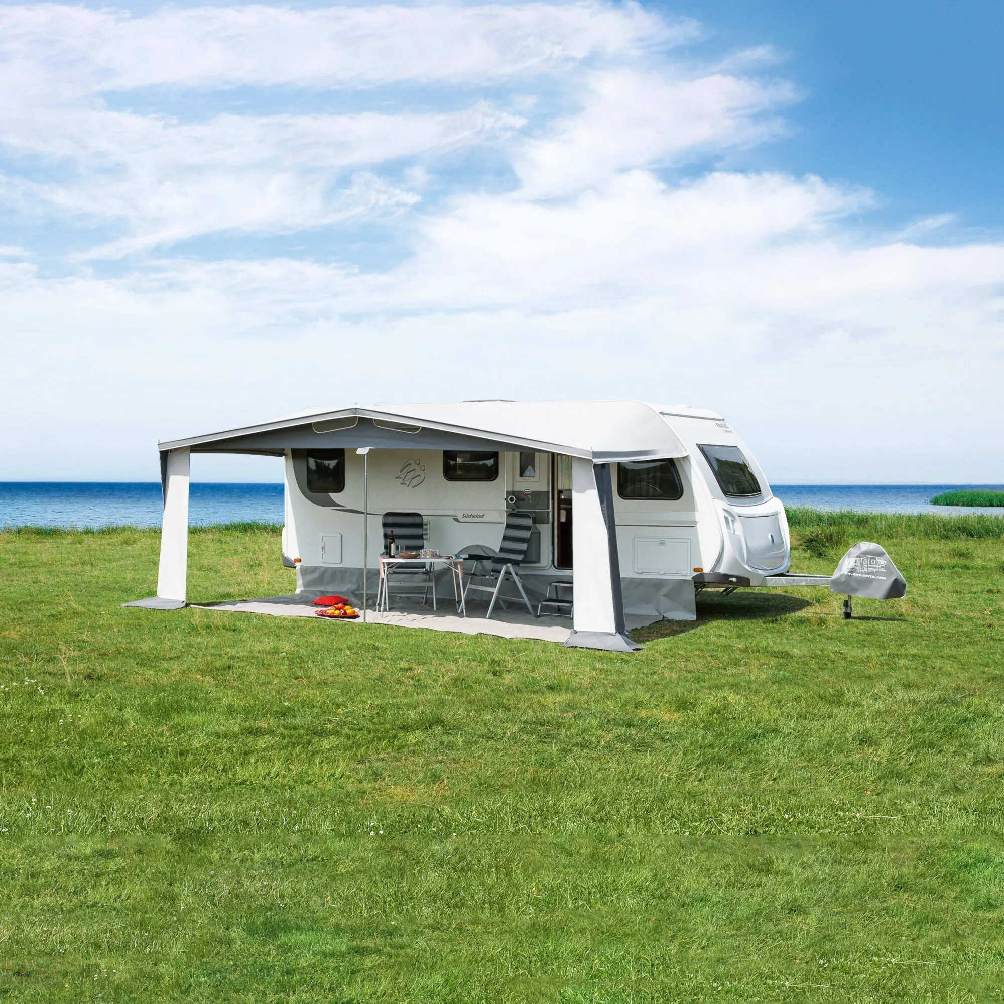 Grauweißes Saisonvorzelt Lux mit 1000 cm Breite an einem Wohnwagen auf einer Wiese am Meer, mit Tisch und Stühlen darunter