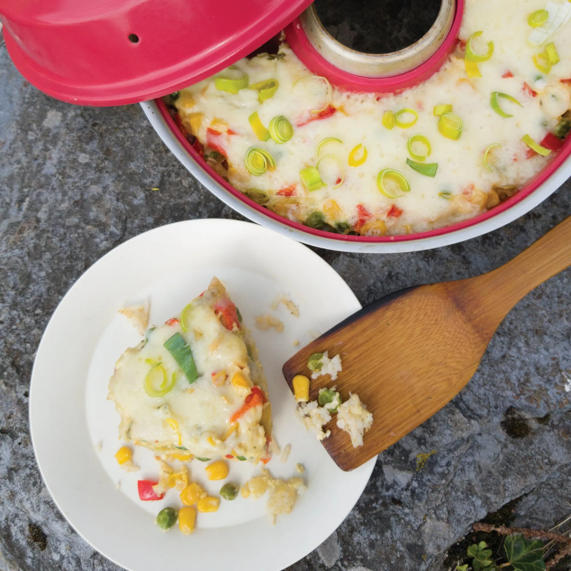 Geöffneter roter Omnia Backofen mit überbackenem Gemüseauflauf, daneben ein Teller mit Portion und Holzspatel