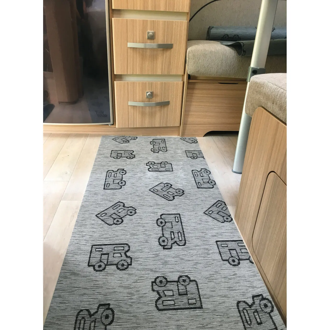 Grauer Läufer mit schwarzen Fahrzeug-Silhouetten liegt im Camper-Innenraum vor Holzmöbeln, Sofa und Schubladen