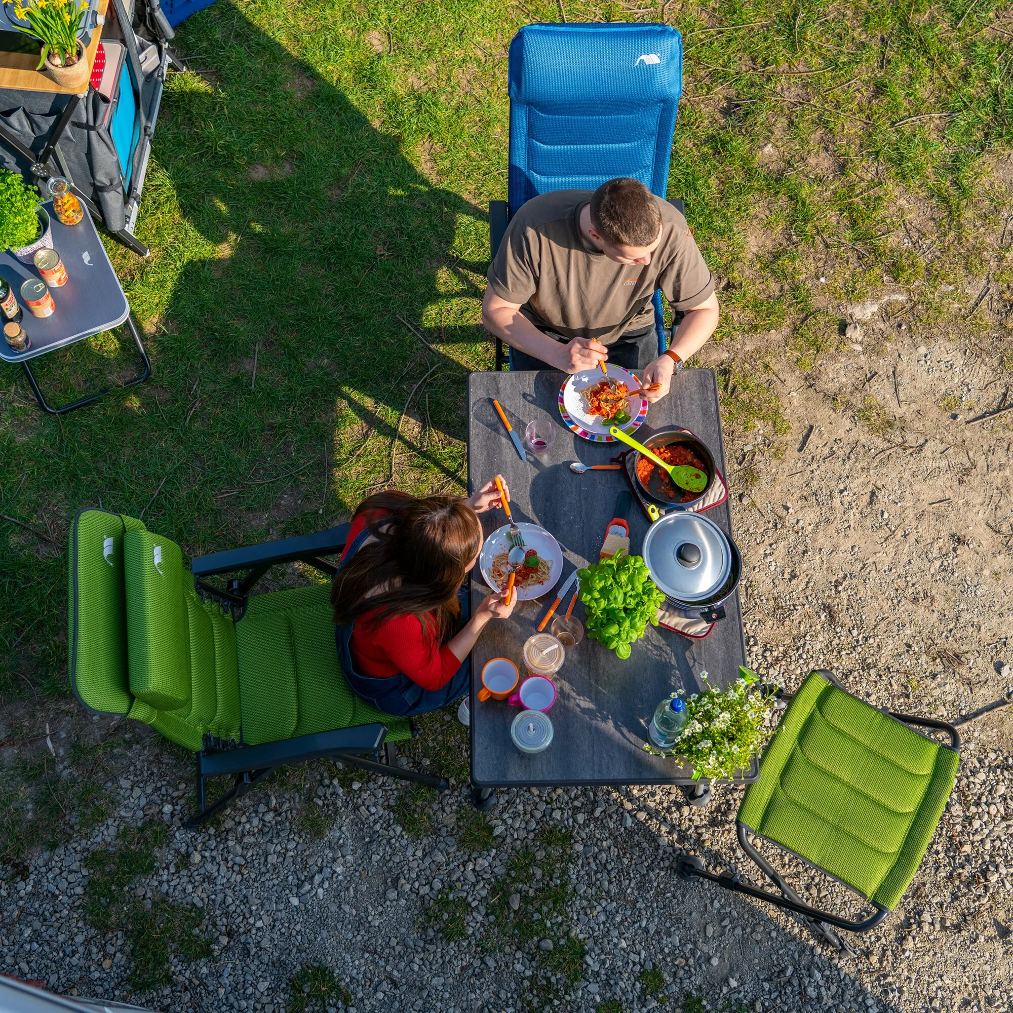 Schwarzer Campingtisch HighQ Blackline, 115 cm, mit Speisen und Geschirr beim gemeinsamen Essen im Freien окружgeben von Campingstühlen