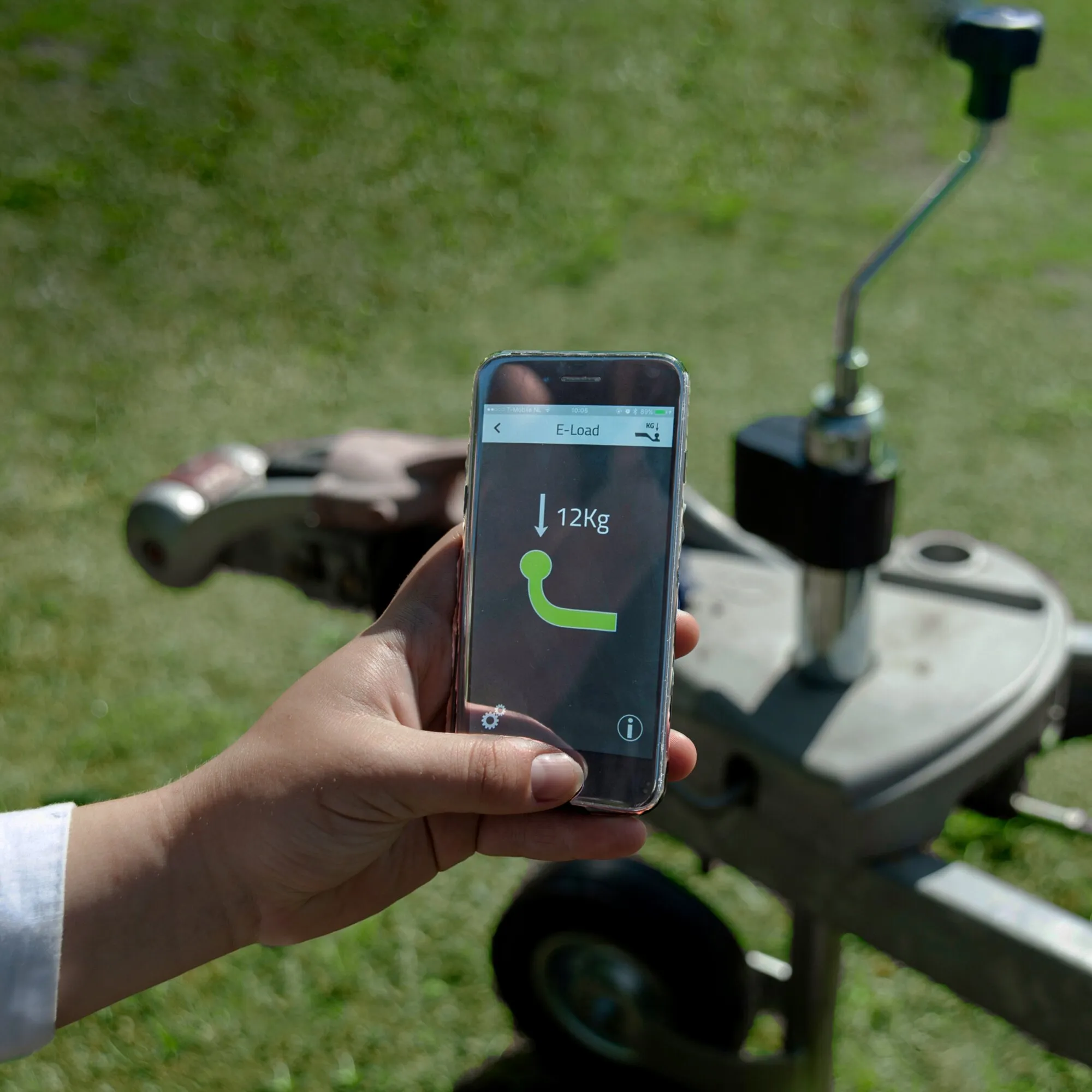 Hand hält ein Smartphone mit E-Load-App und Anzeige von 12 kg neben einem Stützlastsensor an einer Anhängerkupplung im Freien