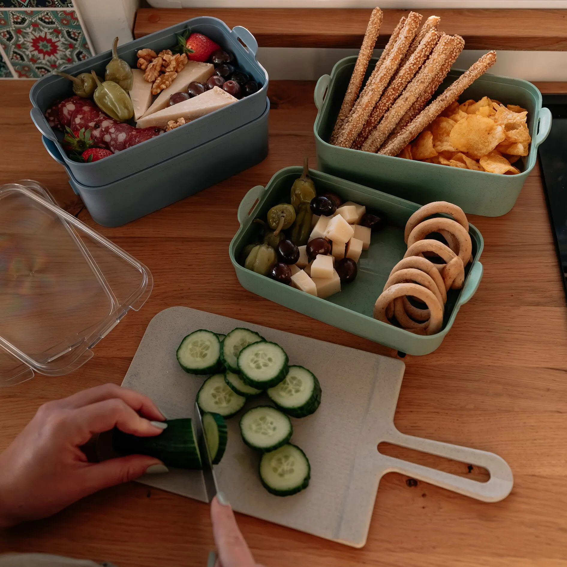 Grüne STAKK BentoBox 2 mit mehreren Fächern, Sesamstangen und Chips, Käsewürfel, Oliven und Trauben; Vordergrund Gurkenscheiben auf Schneidbrett